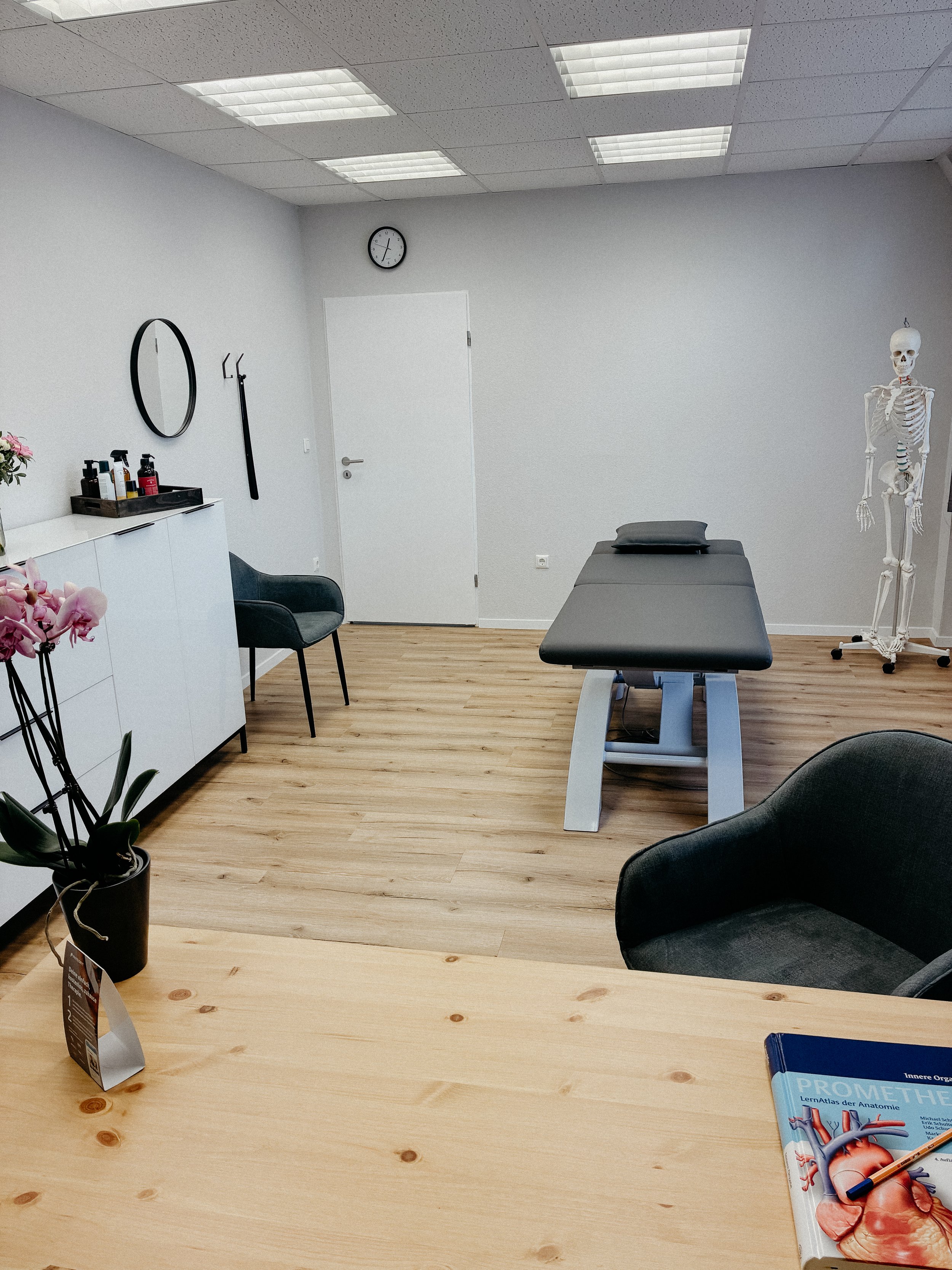 Ein Behandlungsraum mit einem Behandlungsbett, einem Stuhl, einem Schrank mit Pflegemitteln, einem Spiegel, einer Pflanze, einer Anatomie-Schädelmodell und einem Buch über die Anatomie des Herzens.