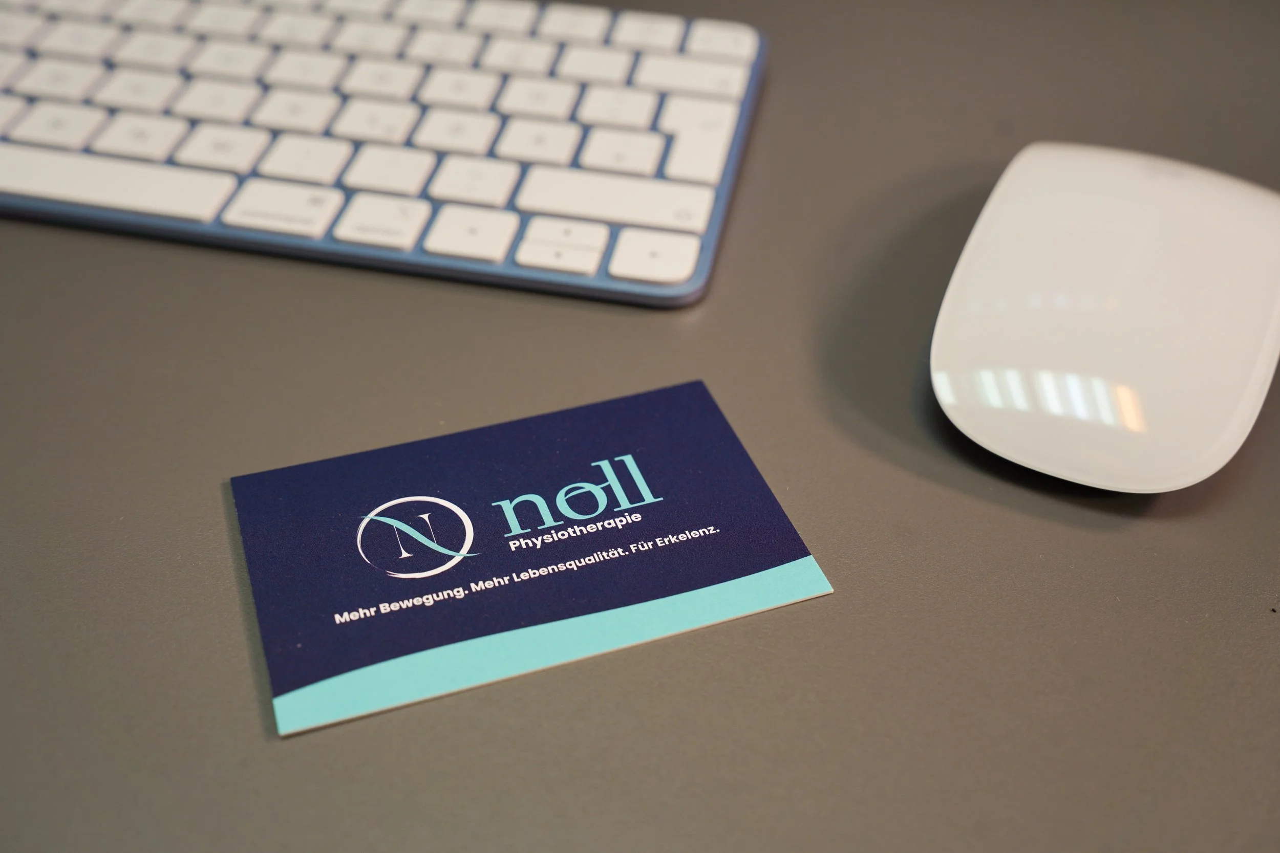 Eine Visitenkarte für eine Physiotherapie namens 'nall' liegt auf einem grauen Tisch neben einer weißen Tastatur und einer weißen Computermaus.