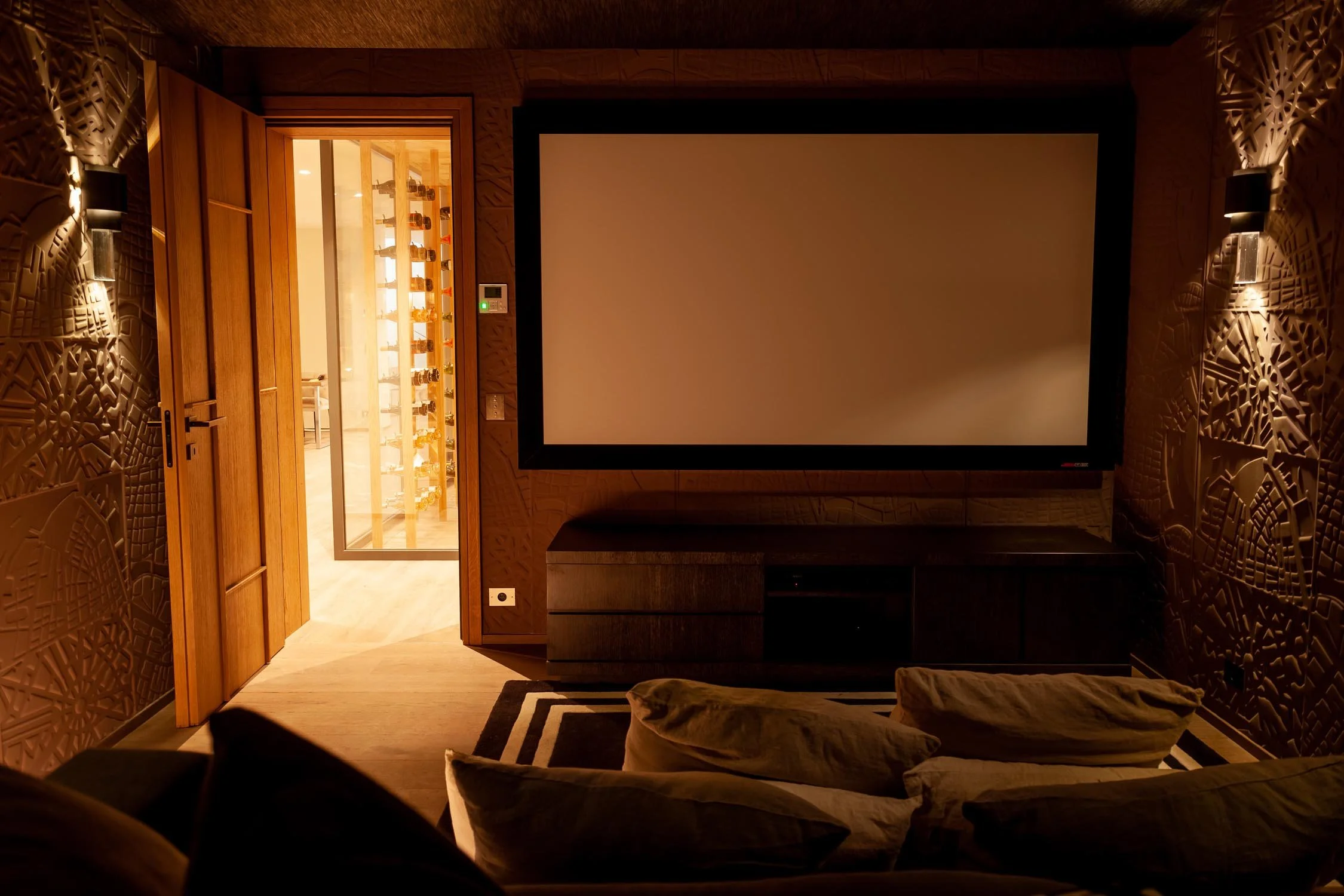 Salon avec un grand écran de télévision, mur en bois avec décorations en relief, canapés avec coussins, porte en bois menant à une pièce avec étagère contenant des bouteilles, ambiance chaleureuse et tamisée.