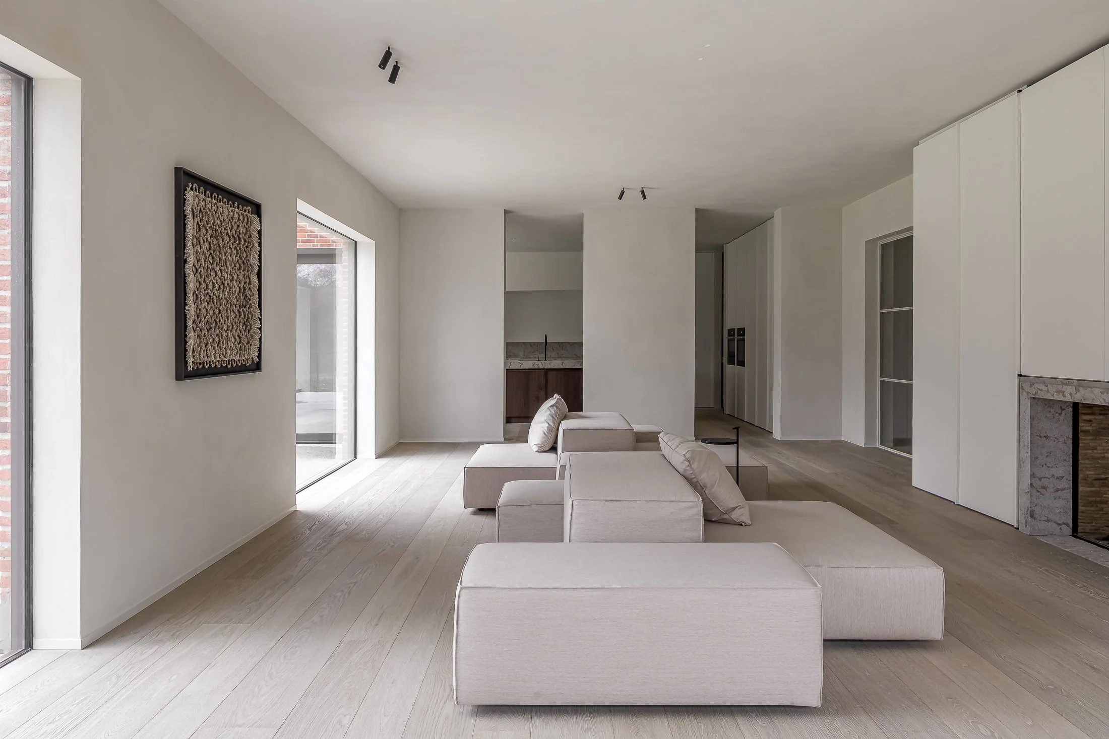 Intérieur d'un salon moderne avec sofa beige, murs blancs, cheminée, et fenêtres en verre laissant entrer beaucoup de lumière