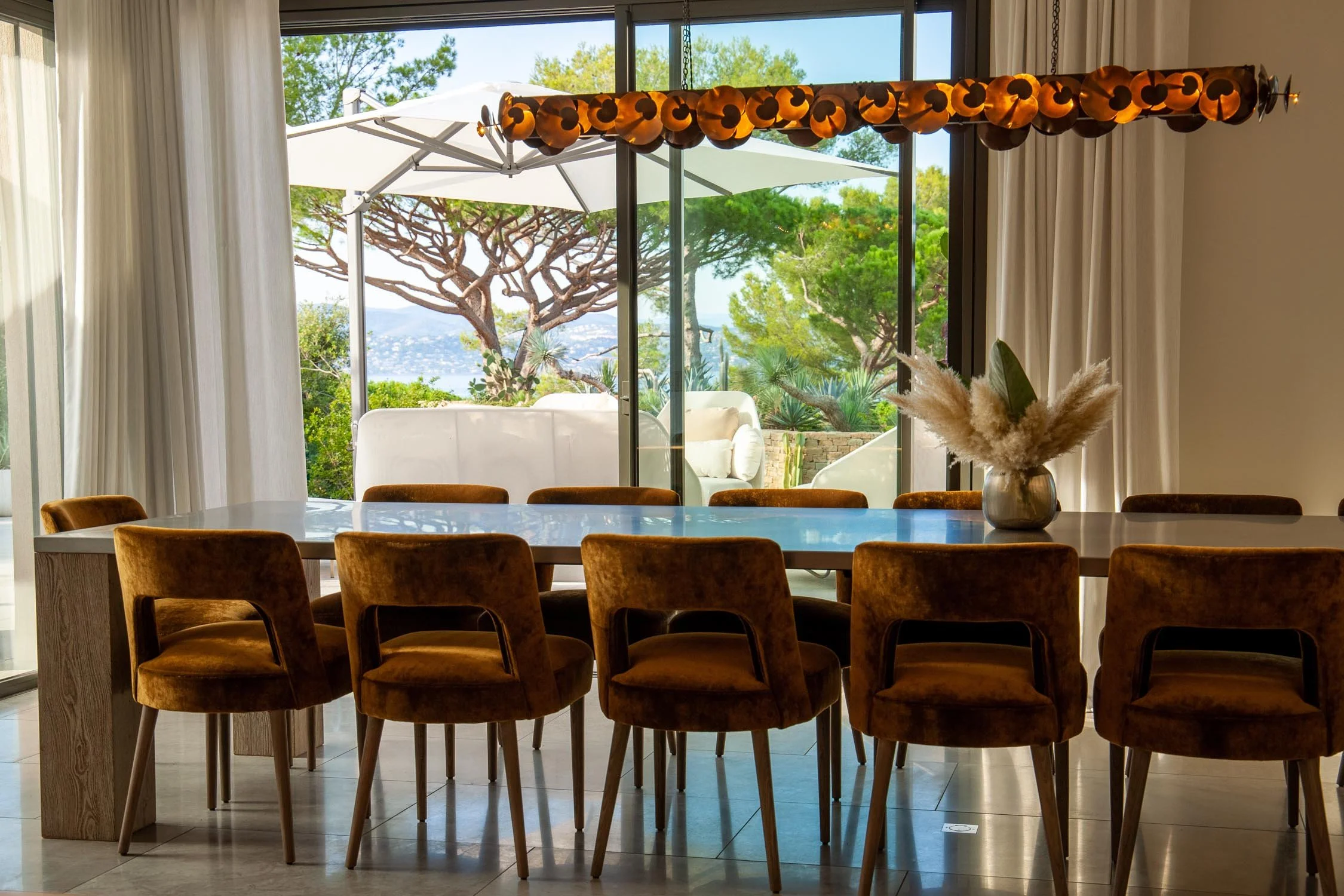 Salle à manger intérieure avec une grande table en bois, plusieurs chaises en velours marron, et un vase avec des fleurs séchées. En arrière-plan, une grande baie vitrée donnant sur un jardin avec des arbres et un parasol blanc.
