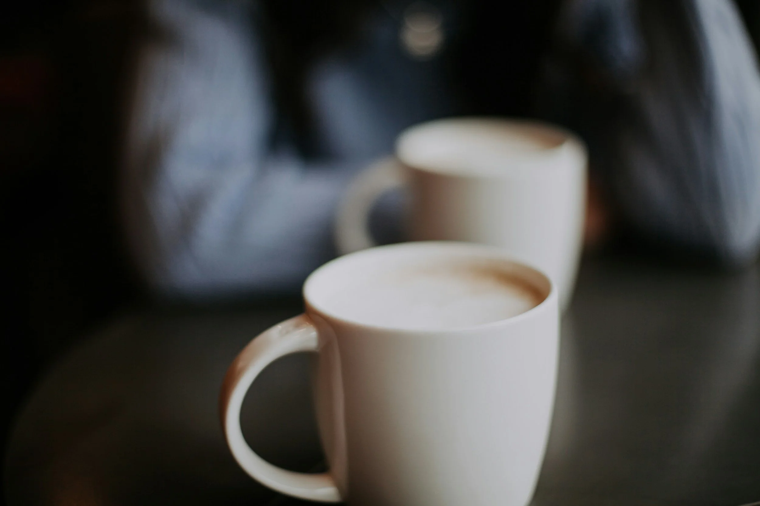 Two white ceramic mugs filled with coffee on a dark surface, with a blurred background.
