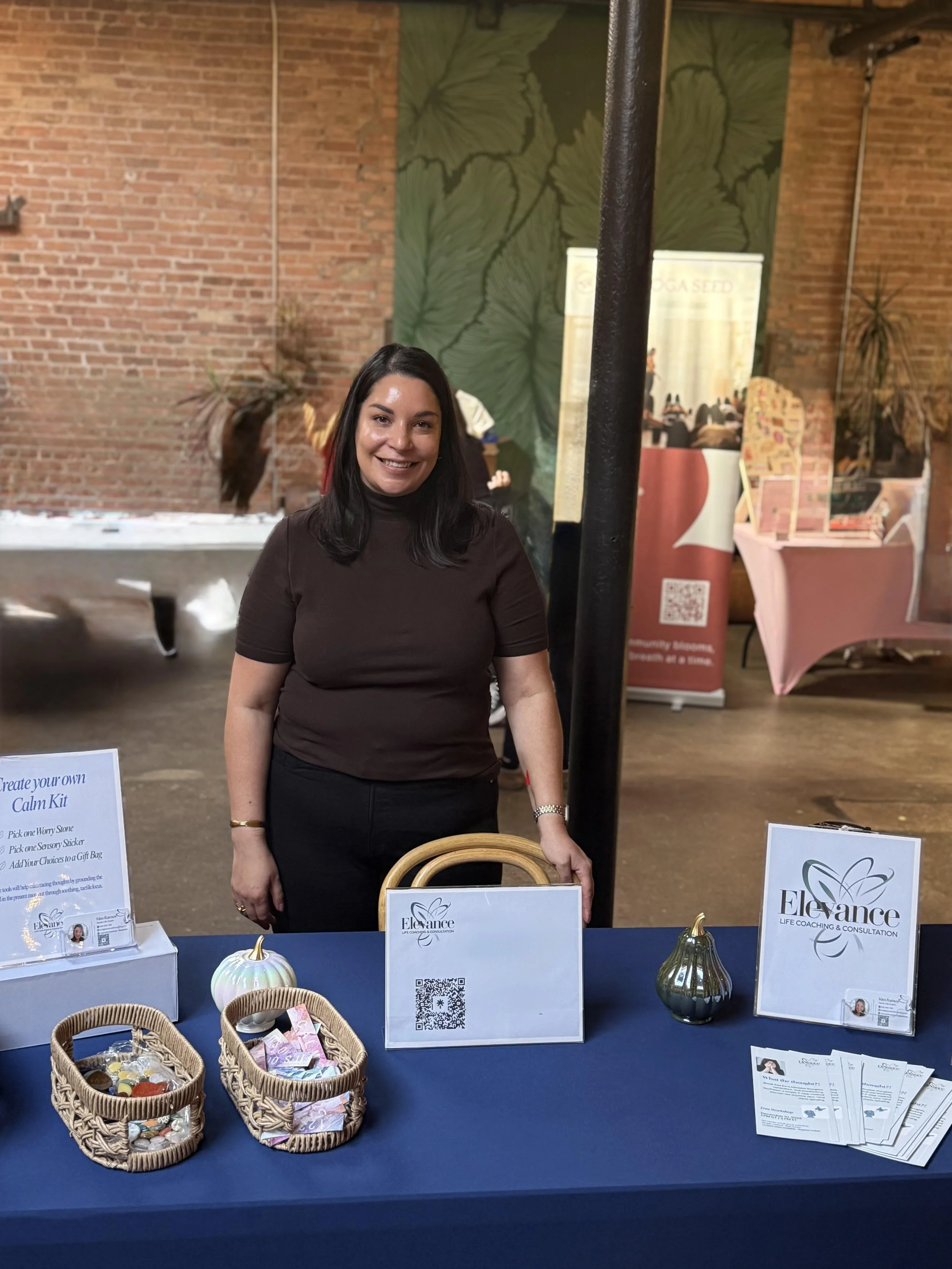Coach Kim at a table with promotional materials for Elevance Life Coaching & Consultation, including baskets of worry stones, a sign, and flyers, indoors with brick walls and plants in the background.