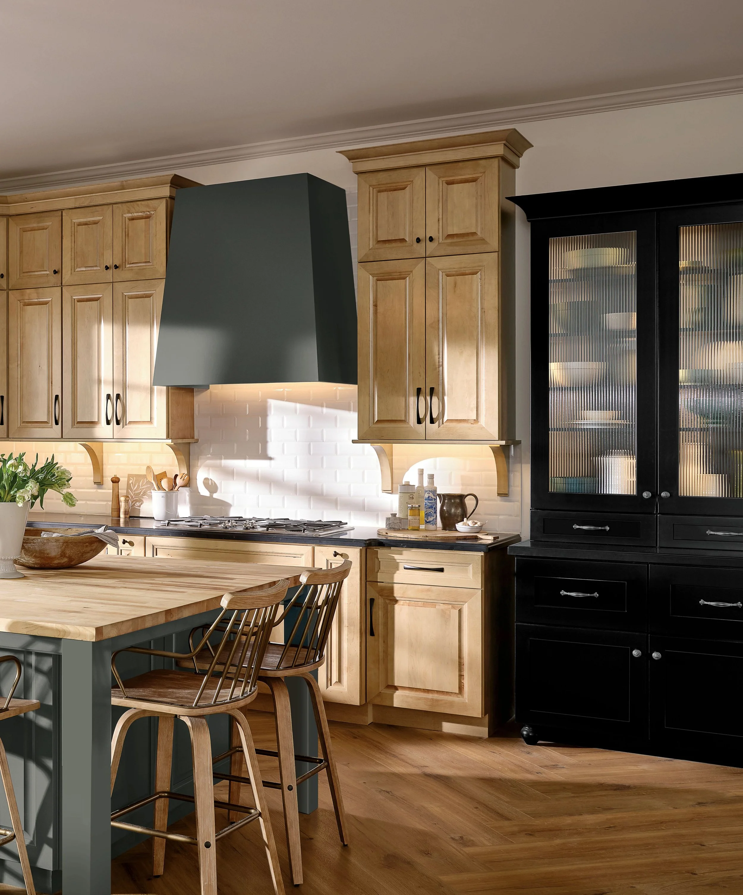 A kitchen with light wooden cabinets, black handles, a black hutch, and a large gray range hood over a stove. There's a kitchen island with a light wood top and wooden and black metal chairs, with various jars and kitchenware on the counters.