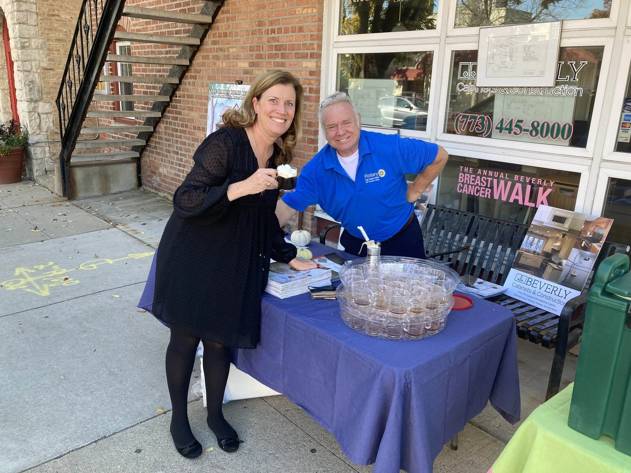 A smiling woman with light brown hair in a black dress and black shoes holding a cup of beverage standing next to a man in a blue polo shirt with Rotary logo, behind a table with clear cups and a purple tablecloth outside a building with business signs.