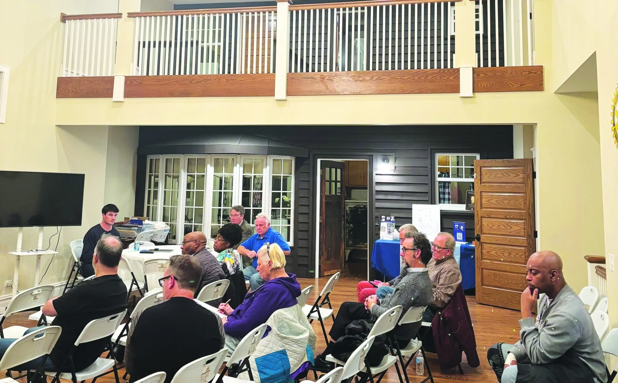 A group of people sitting on white chairs in a room, attending a meeting or presentation, with a man speaking at the front. The room has a second-story balcony, large windows, and wooden flooring.
