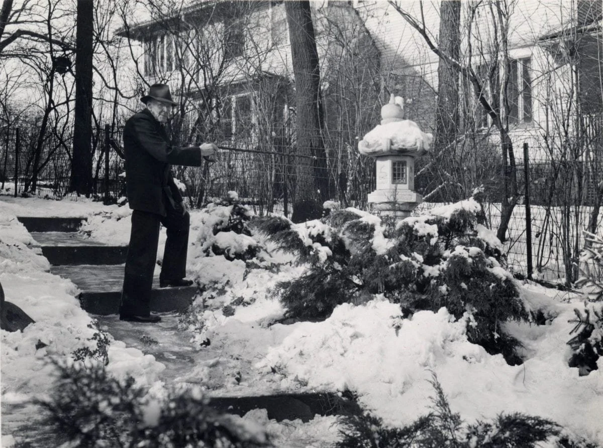 An elderly man in a dark coat and hat is shoveling snow on the steps of a sidewalk, with snow-covered bushes and a traditional lantern in the background.