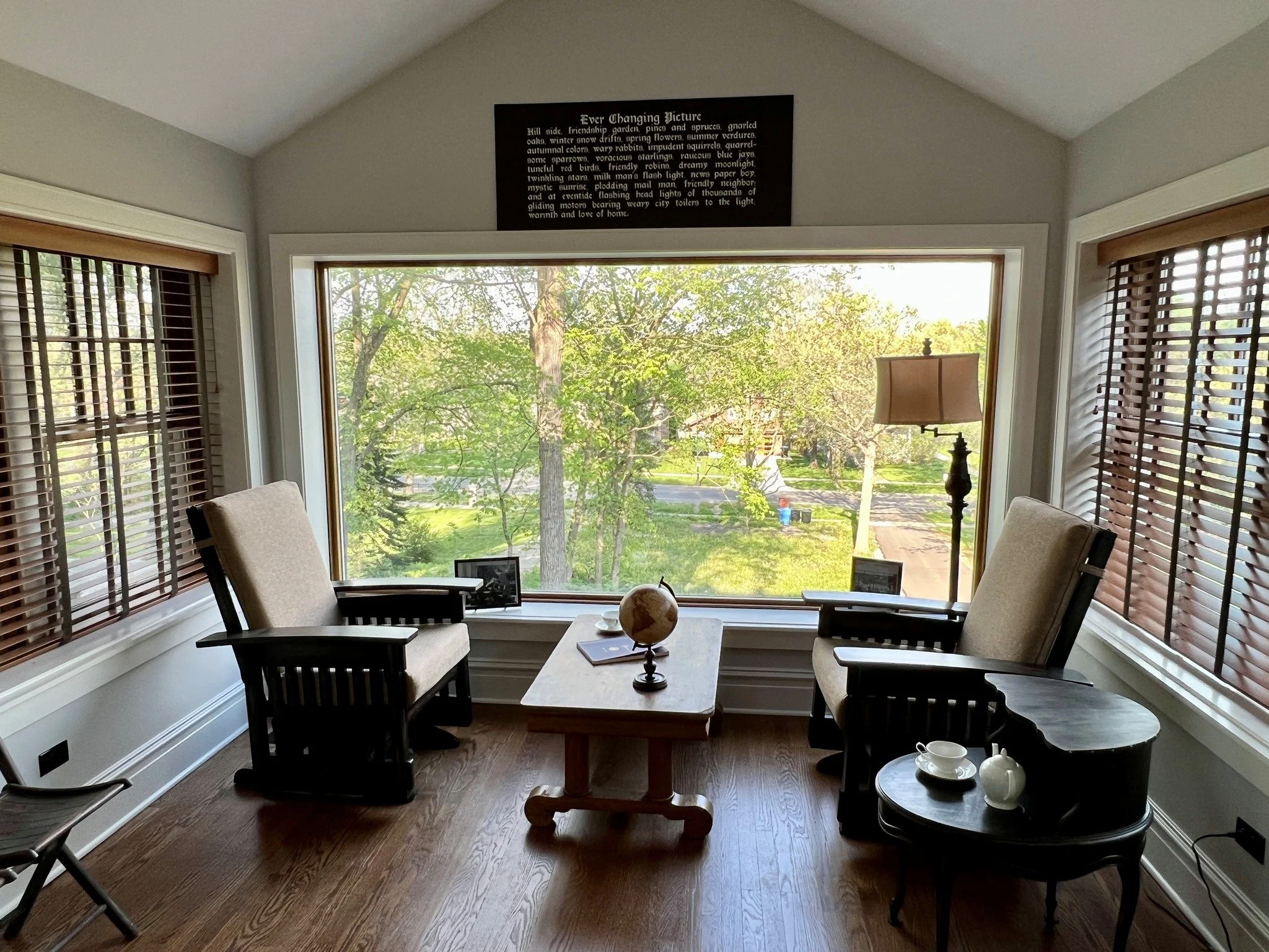 A cozy sitting area with two beige armchairs and a small black side table with a teacup and teapot. A rectangular wooden table with a globe and books sits between a large bay window, which has a view of trees and greenery outside. A tall floor lamp is near the right armchair. A picture frame and a small black object are on the window sill. Above the window, there is a black sign with white text.