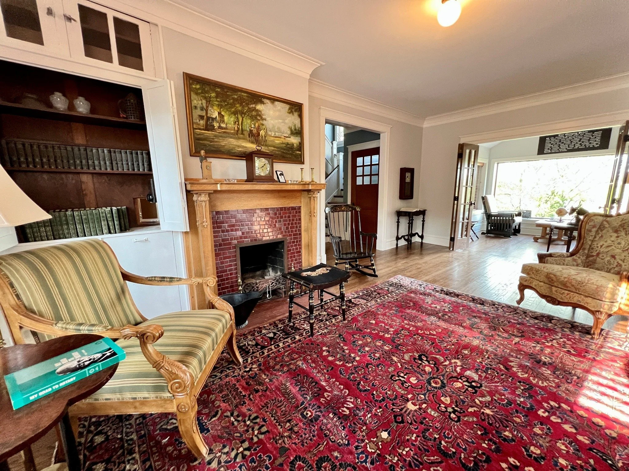 Living room with a fireplace, vintage furniture, built-in bookshelves, a large window, and hardwood floors.