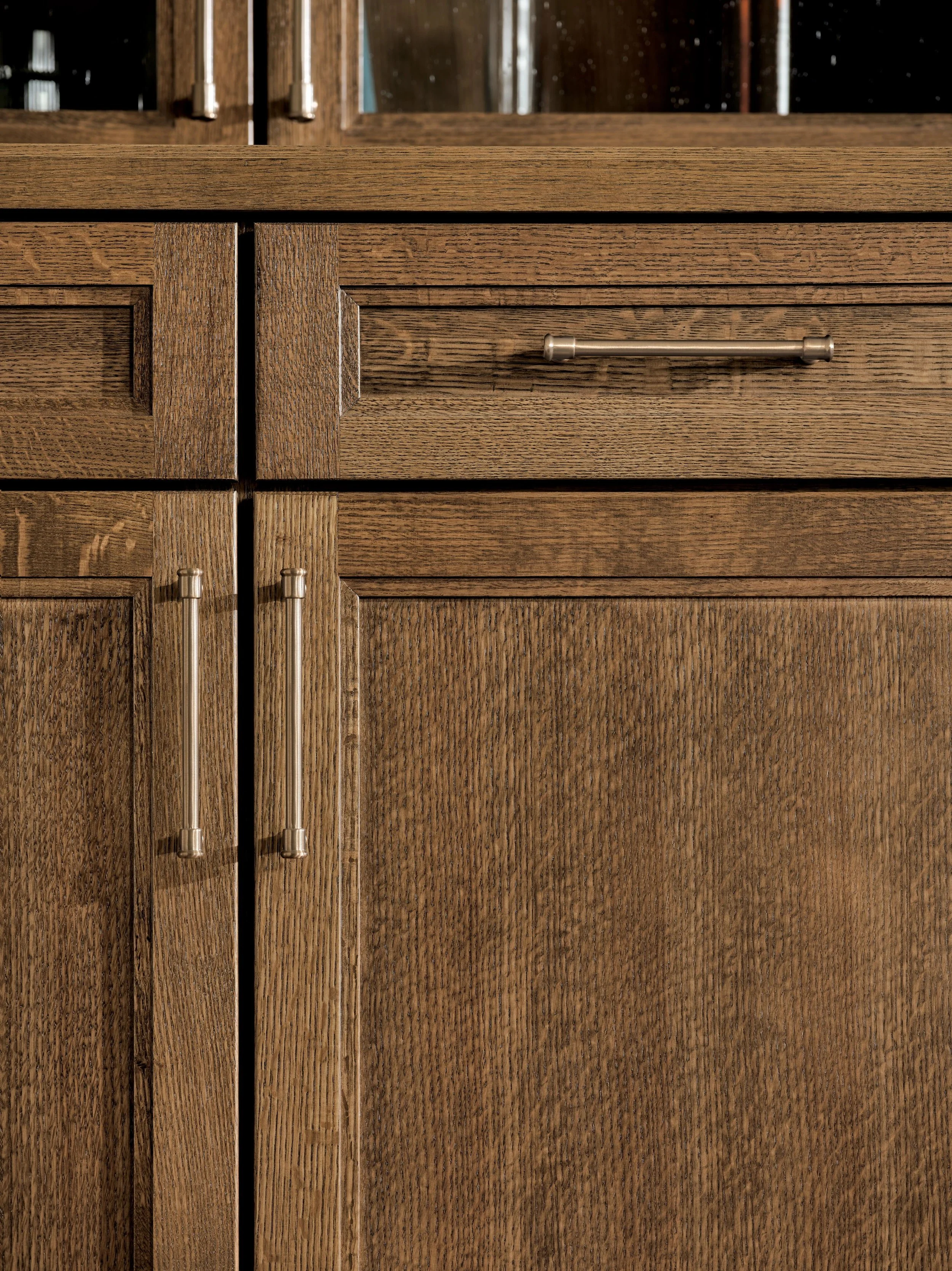 Close-up of a wooden kitchen cabinet with metal handles, showing a glass door at the top with visible glassware inside.