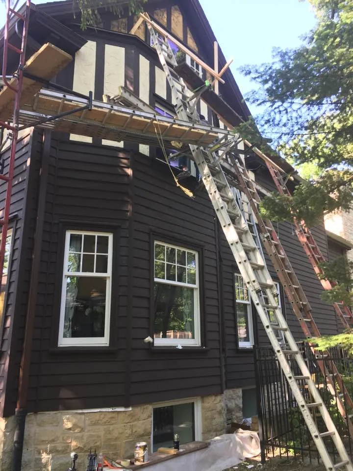 House undergoing exterior siding installation, with multiple ladders and scaffolding in place.