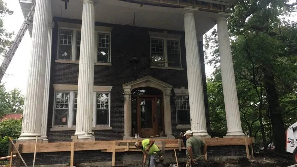 Construction workers installing a new porch or deck in front of a large, historic-style house with tall white columns and brick facade.