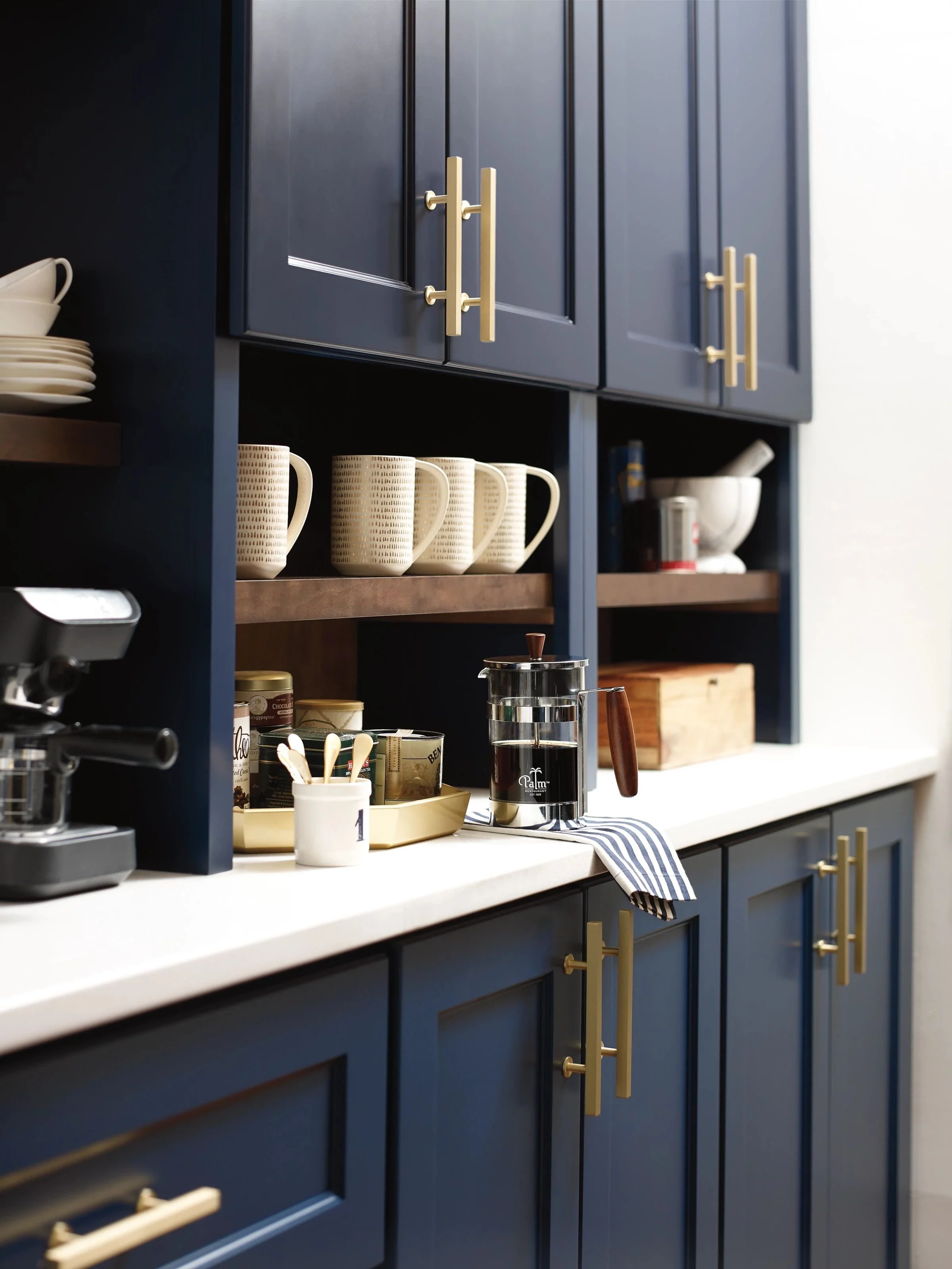 A kitchen with navy blue cabinets featuring gold handles, white countertops, and shelves with beige mugs, white dishes, and various kitchen items, including a French press, spoon holder, and small containers.