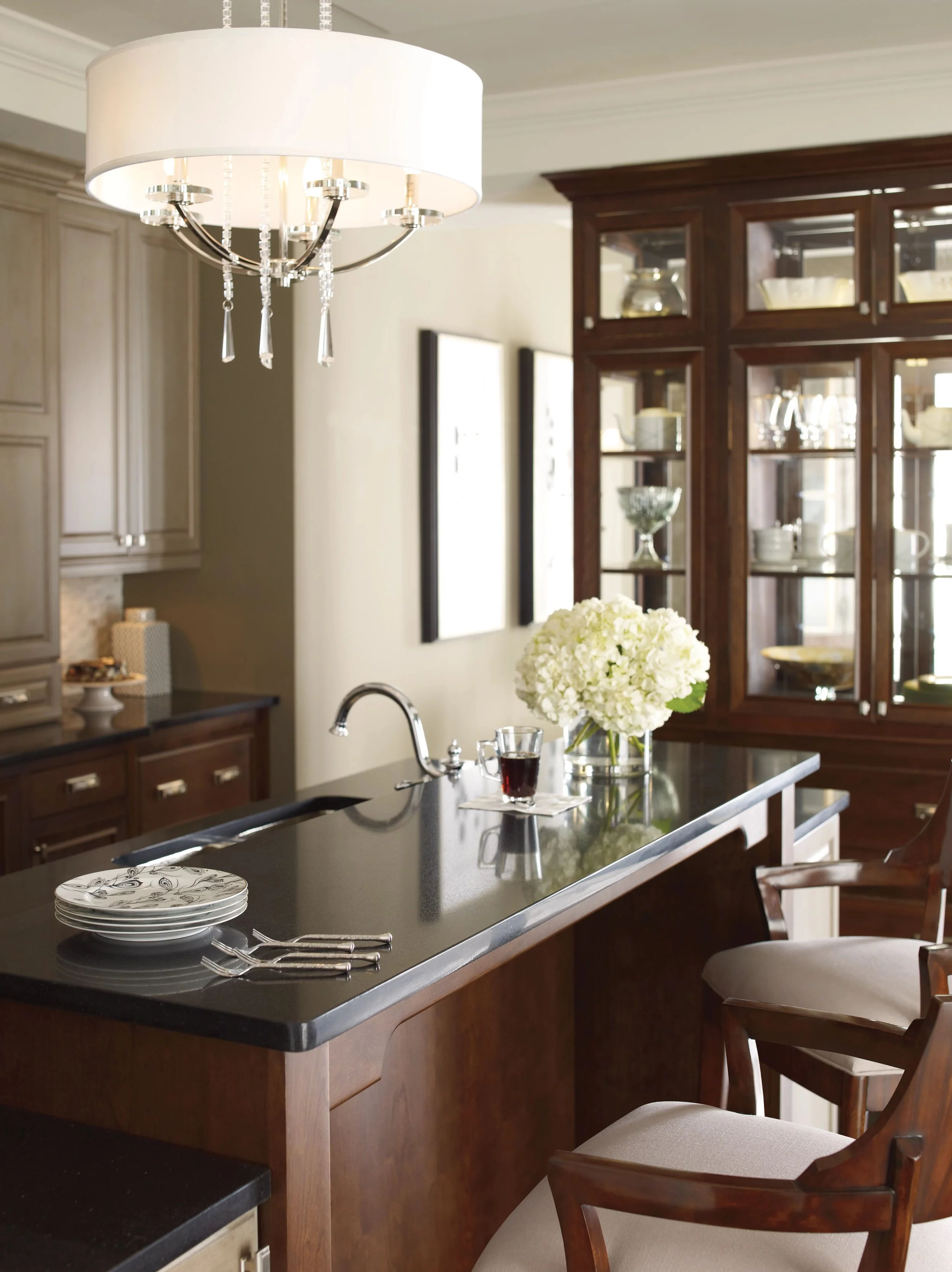 Kitchen with dark wood cabinets, an island with a black countertop, a chandelier, a vase of white flowers, and a plate and cutlery on the counter.