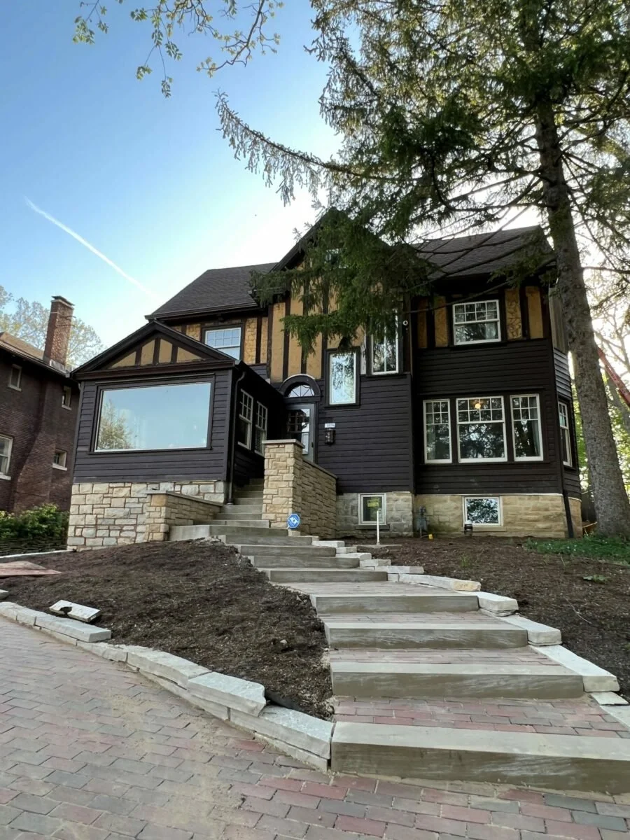 A two-story house under renovation with a black wooden exterior, large windows, stone foundation, and a newly paved concrete staircase leading to the entrance, surrounded by trees and a clear sky.