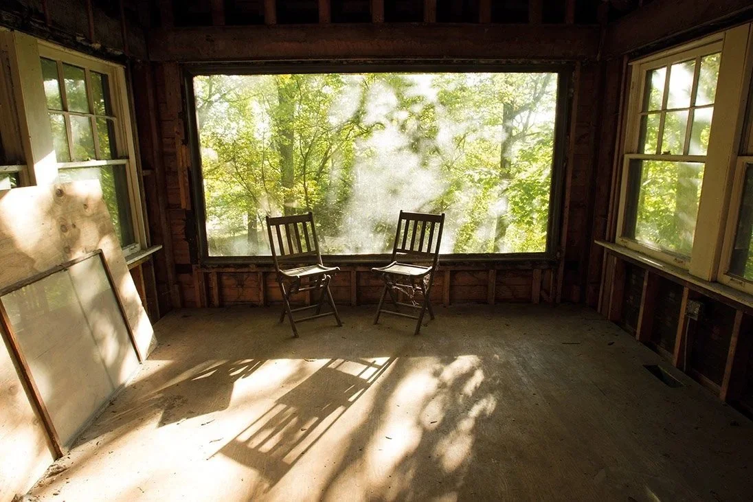 An unfinished room with large windows showing a wooded outdoor scene, two wooden chairs, and sunlight casting shadows on the floor.