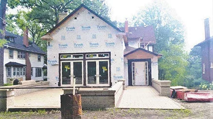 Construction site of a house with a white exterior and large windows, surrounded by trees.
