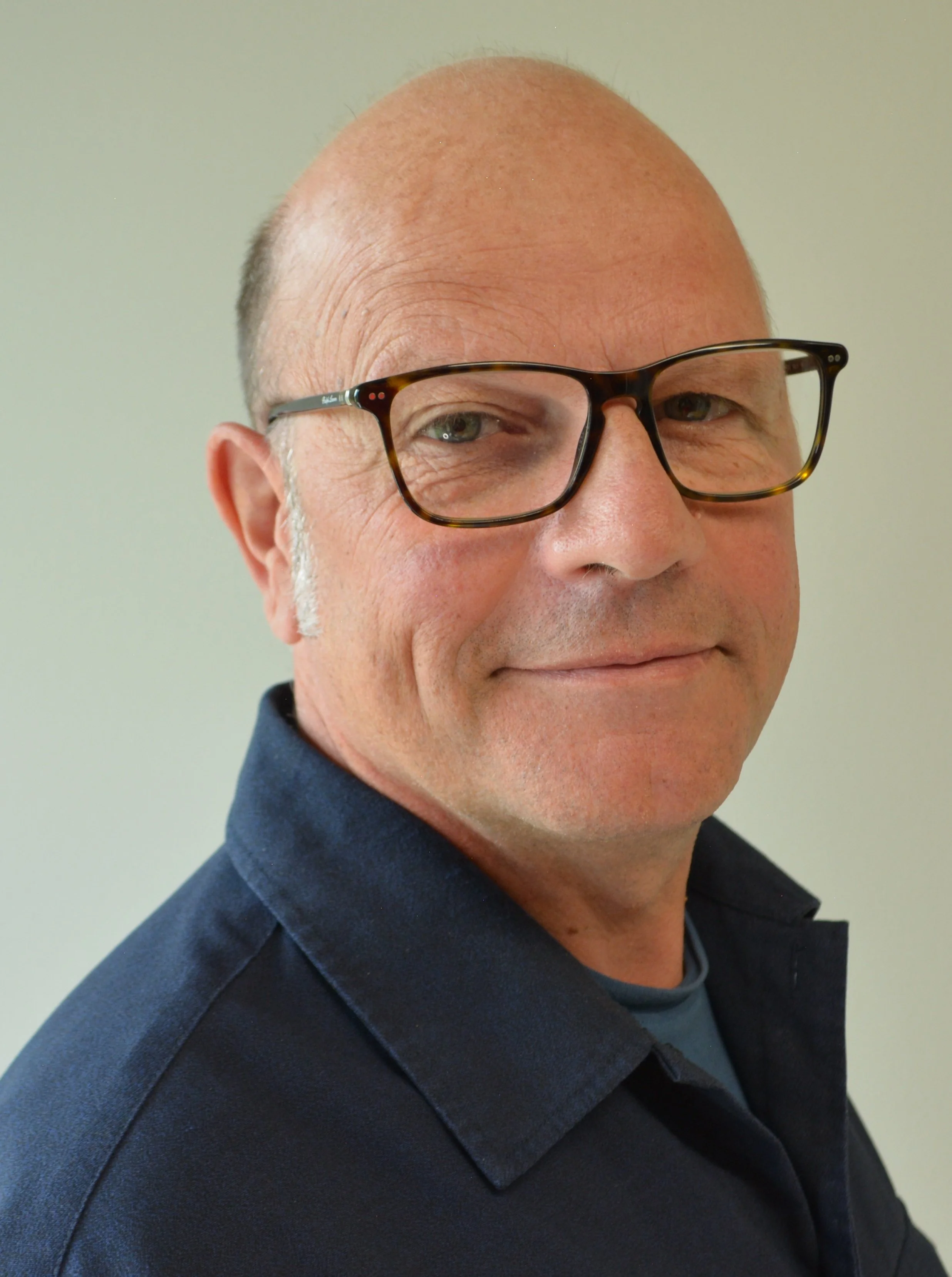 Porträt eines lachenden Mannes mit Gläsern, braun-gestreiften Brillenrahmen, kurzen Haaren und einem blauen Hemd vor hellem Hintergrund.