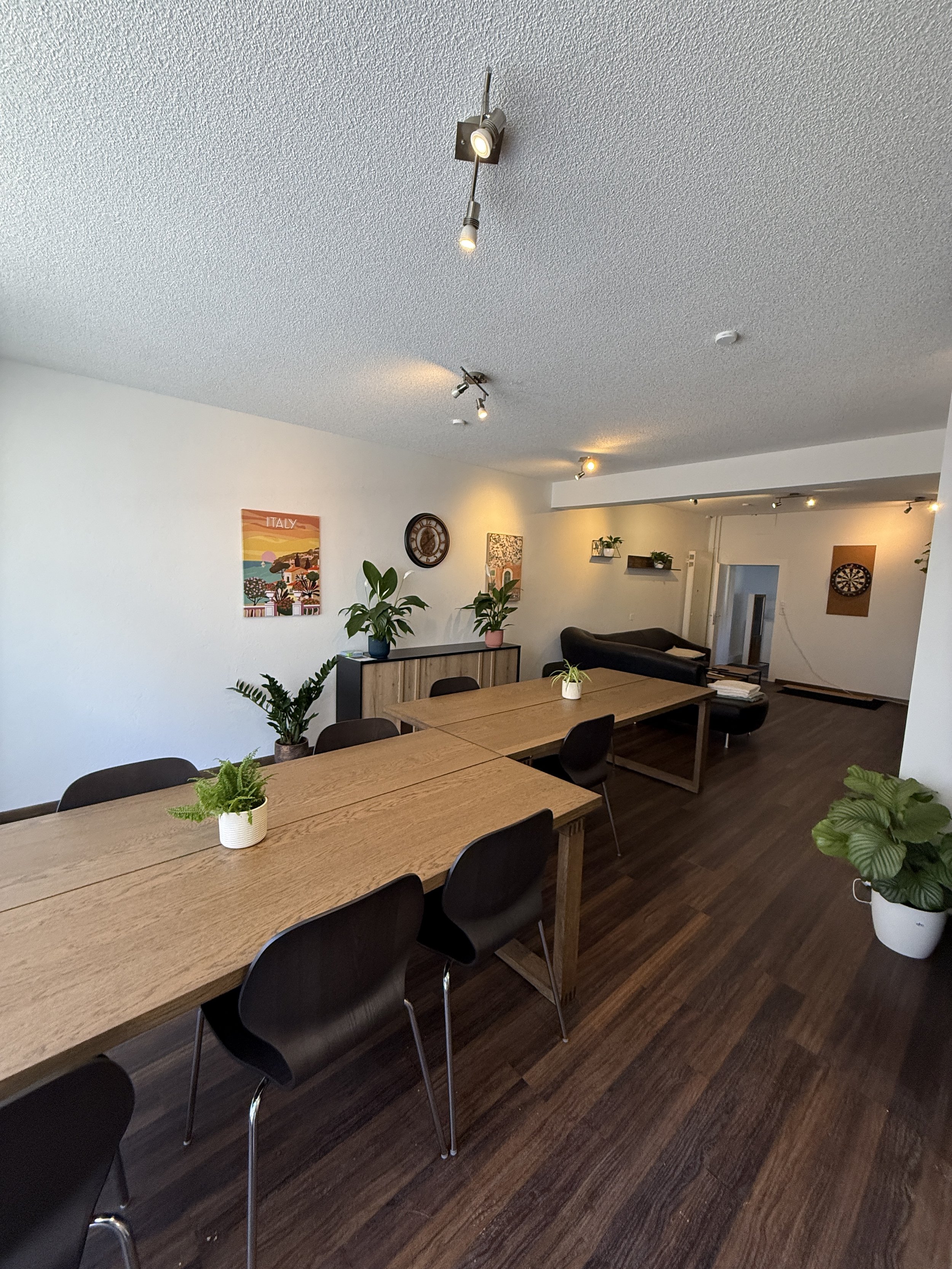 Ein heller Raum mit Holztischen, schwarzen Stühlen, Pflanzen, Wanddekorationen und einem Dartboard an der Wand