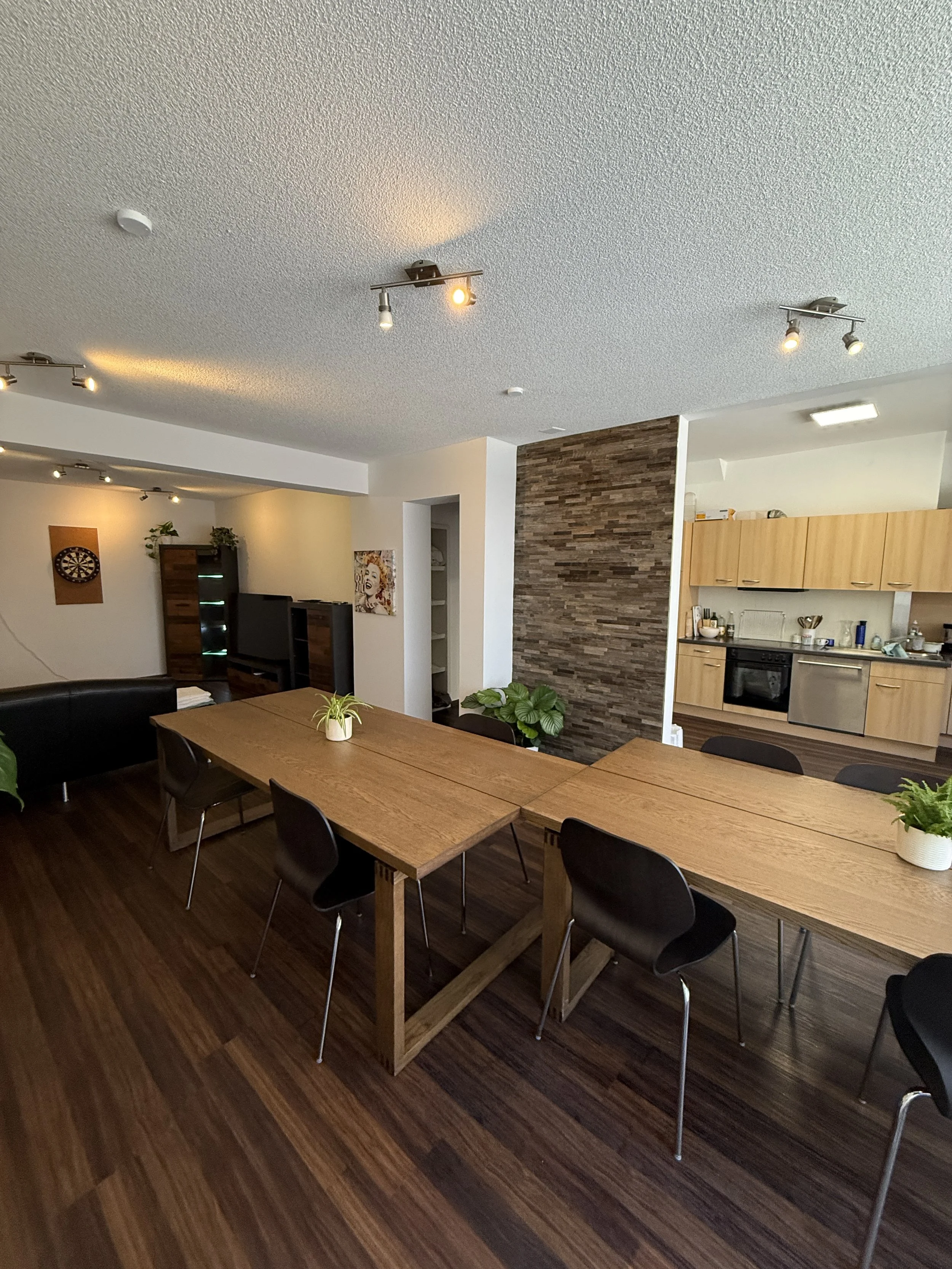 Innenaufnahme einer offenen Wohnküche mit Holztischen, schwarzen Stühlen, Pflanzen, moderner Dekoration und Küchenbereich mit Holzfronten.