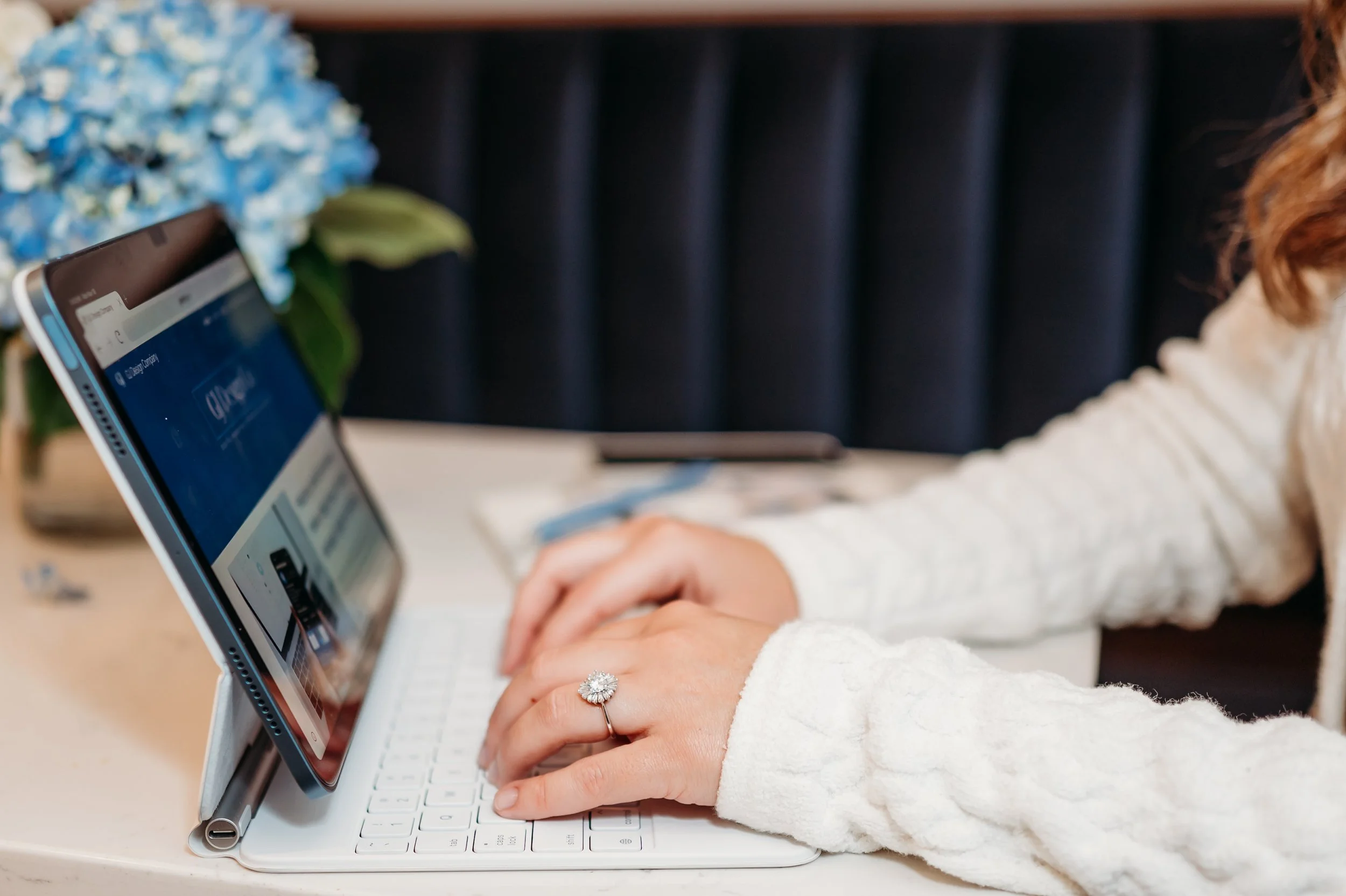 GJ Design Co, a small business digital marketing strategist, typing on a laptop at a desk with a tablet propped up beside the laptop, a vase of blue flowers in the background.