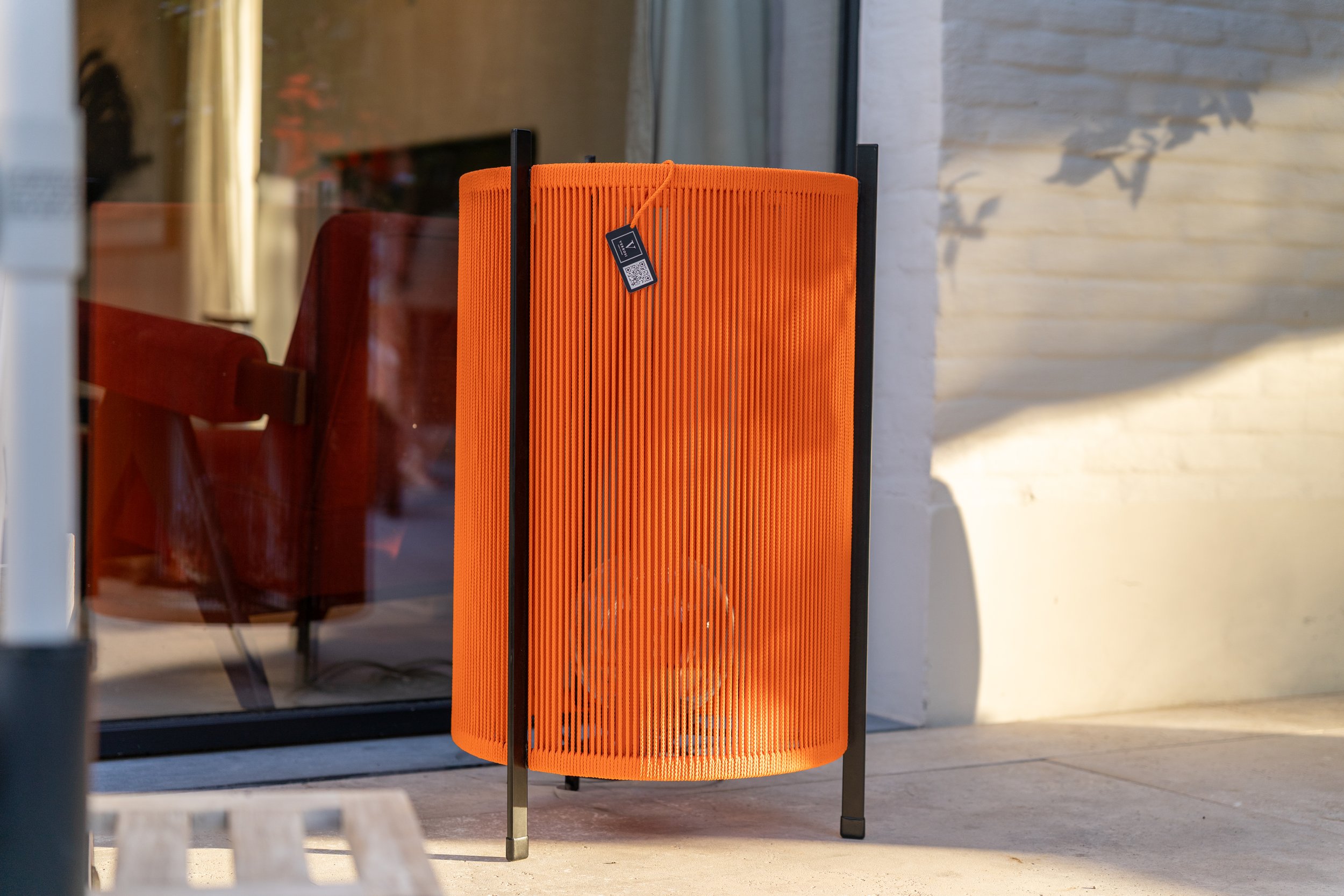 Bright orange cylindrical outdoor lamp with black metal stand, placed on a concrete surface, with a glass door and a beige textured wall in the background.