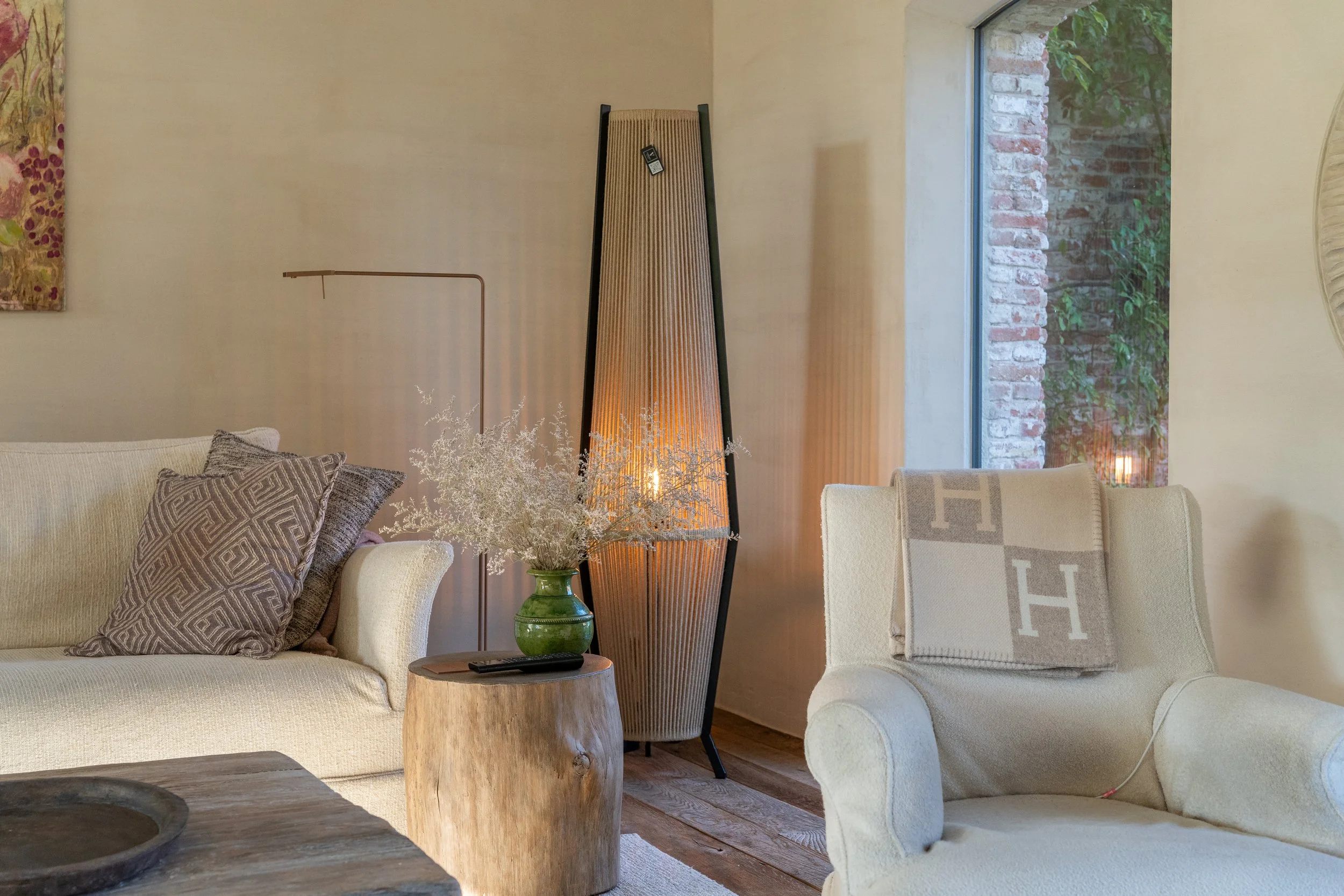 Living room with beige sofa, decorative pillows, wooden side table with green vase and dried flowers, floor lamp, and a large window showing an outside brick wall and green foliage.