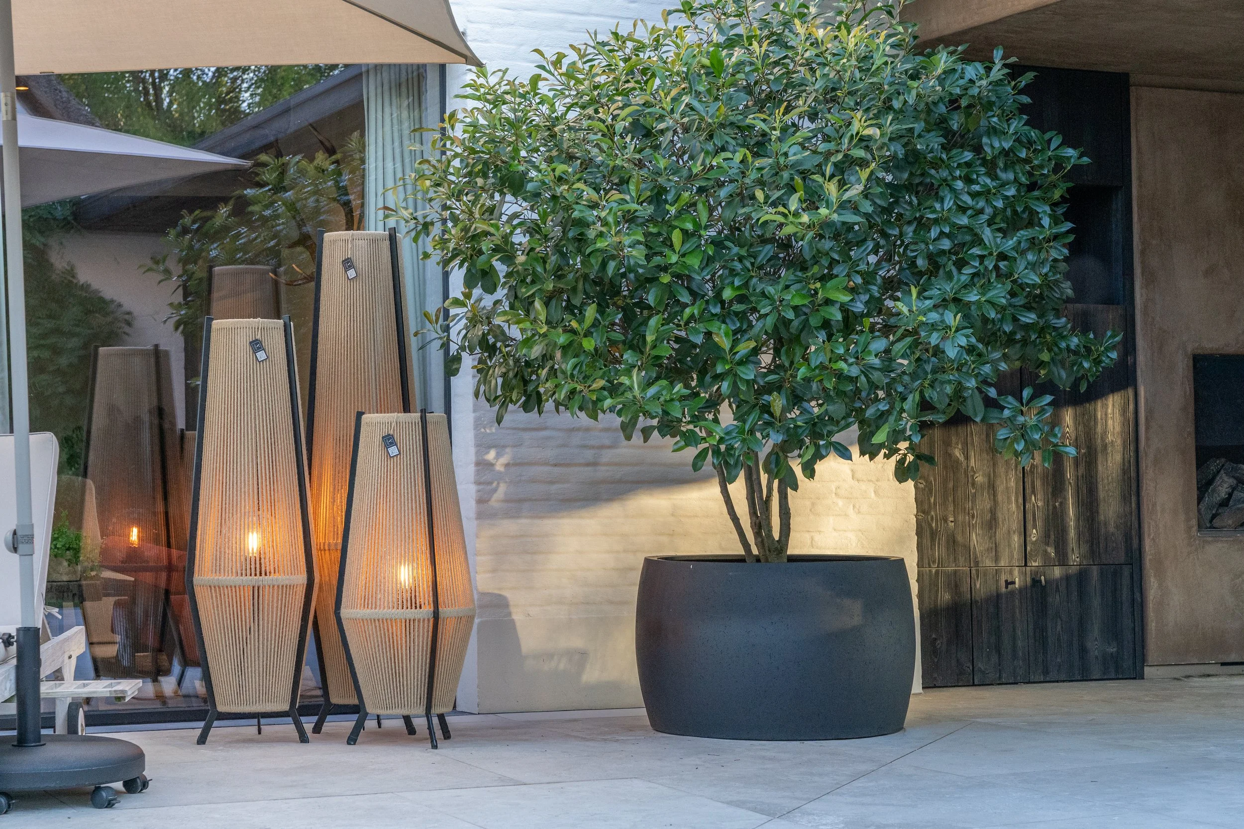 An outdoor patio scene with three tall, wicker-style outdoor heaters with glowing flames near the bottom. A large potted shrub with green leaves is positioned against a beige wall, and a glass window reflects nearby trees and sky. There are umbrellas