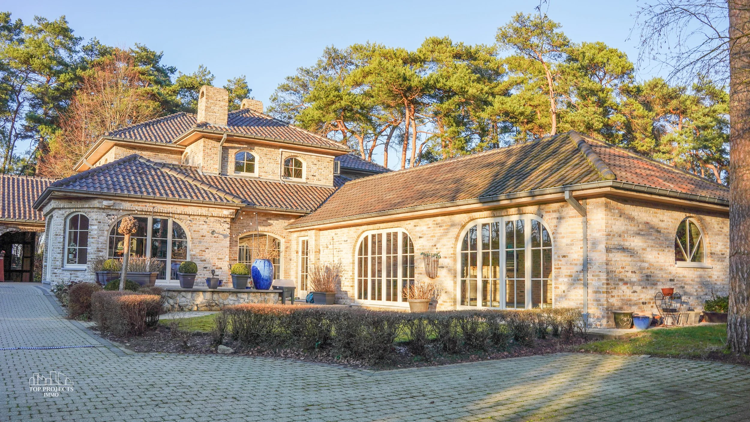 Large brick house with multiple rooflines and large windows, surrounded by trees and a paved driveway