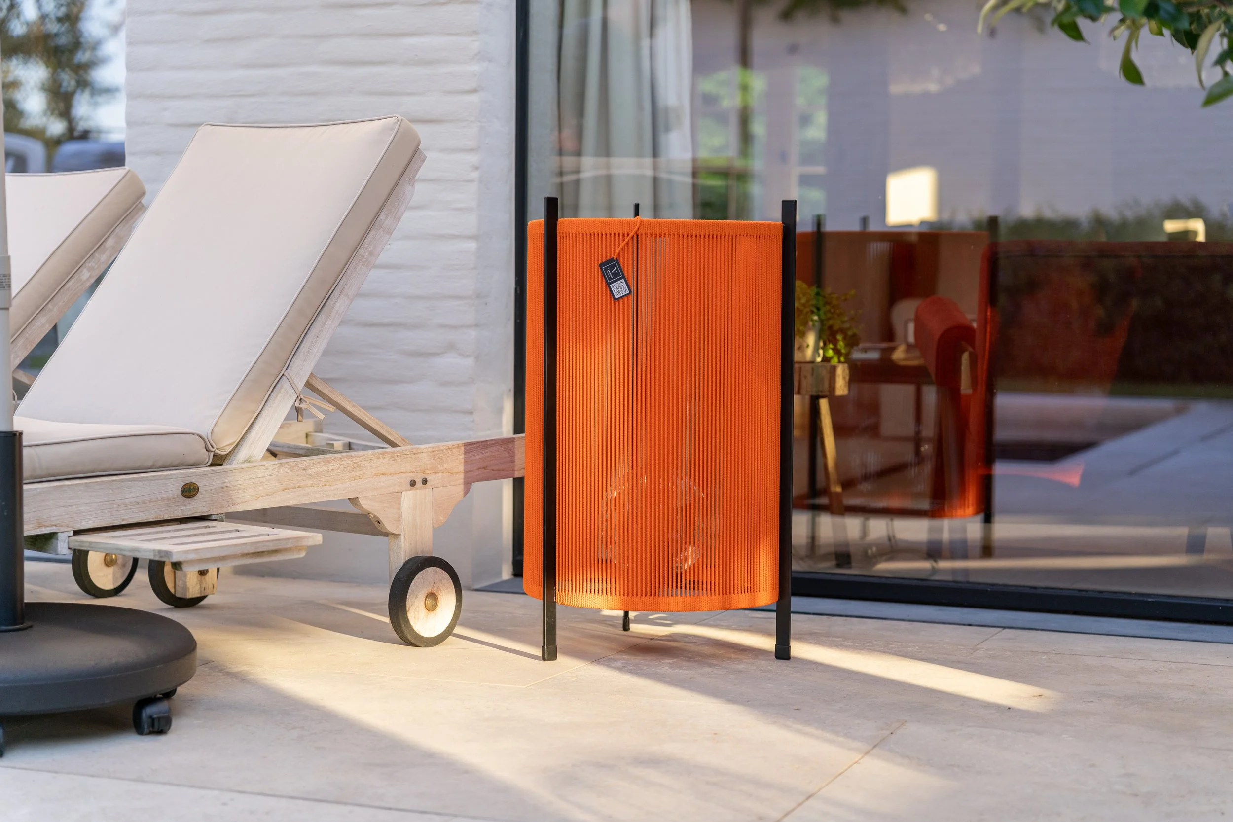 Outdoor patio with white lounge chair on wooden wheelbase, black base, and a bright orange folding screen with QR code tag, reflected interior with red chairs and greenery outside.