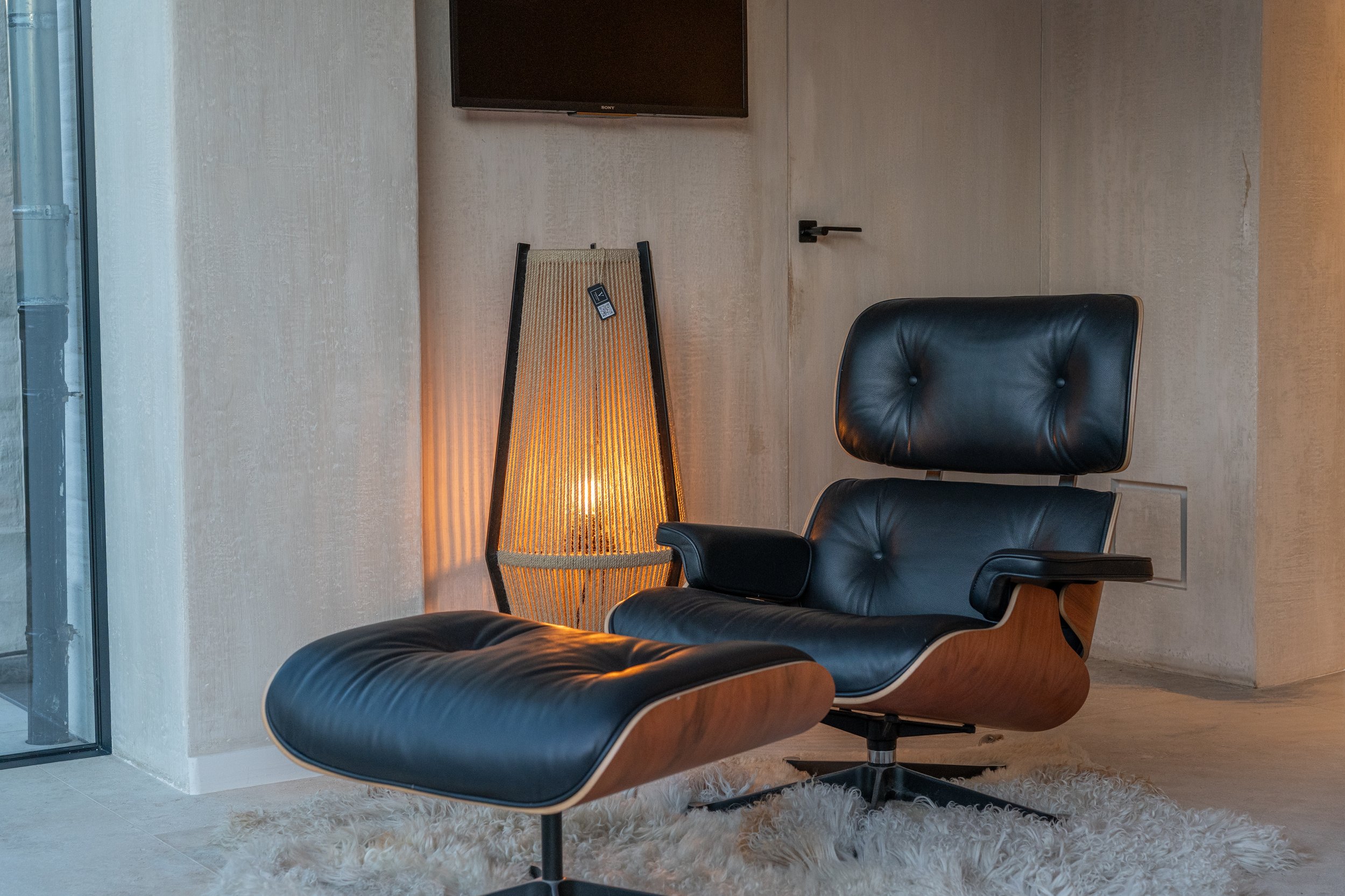 A modern office space with black leather chairs, a tall wooden floor lamp with a warm glow, a cream-colored shag rug, beige textured walls, and a flat-screen TV mounted on the wall.