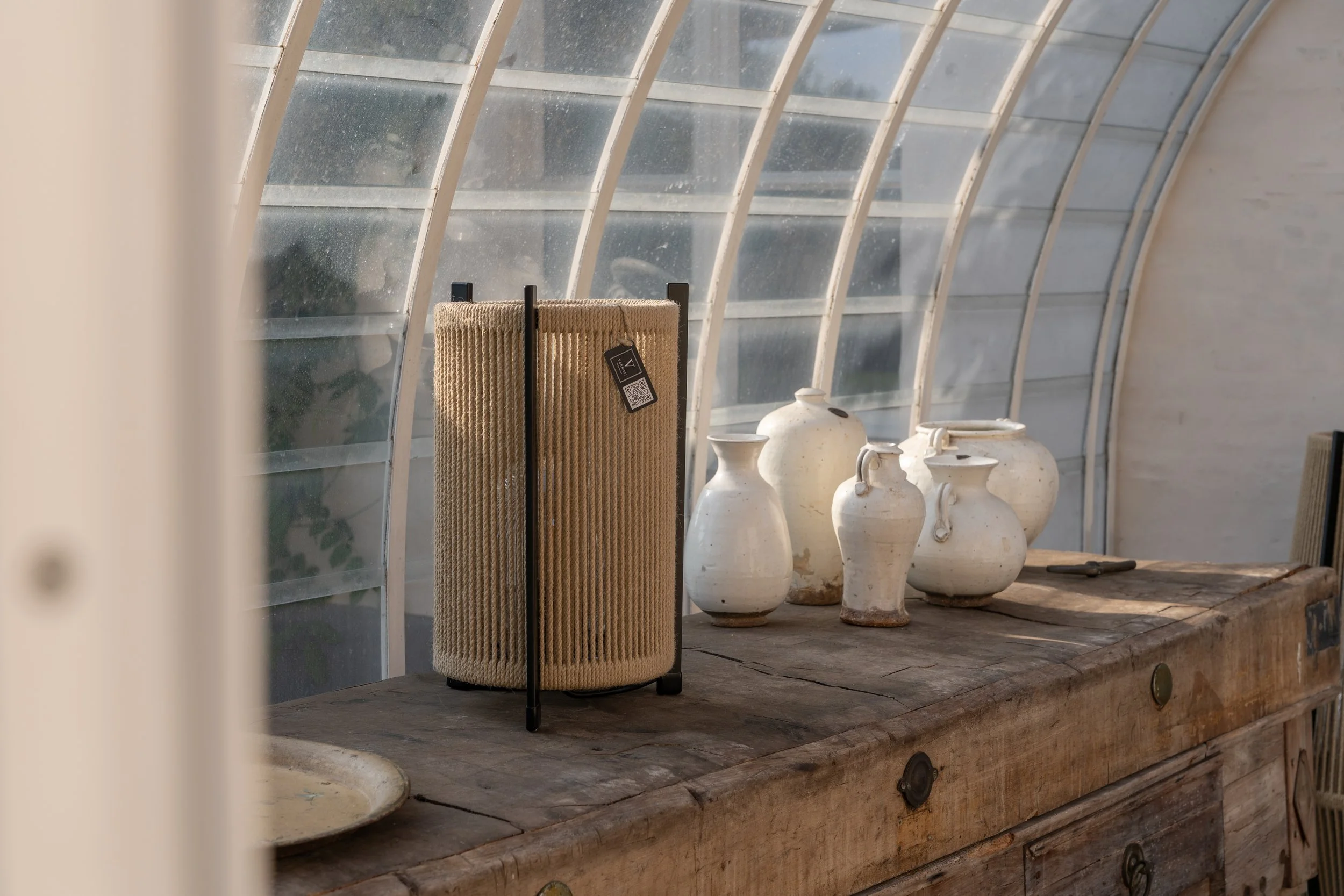 A collection of white ceramic vases and a woven beige standing mirror on a wooden table inside a greenhouse or conservatory with a curved glass roof.