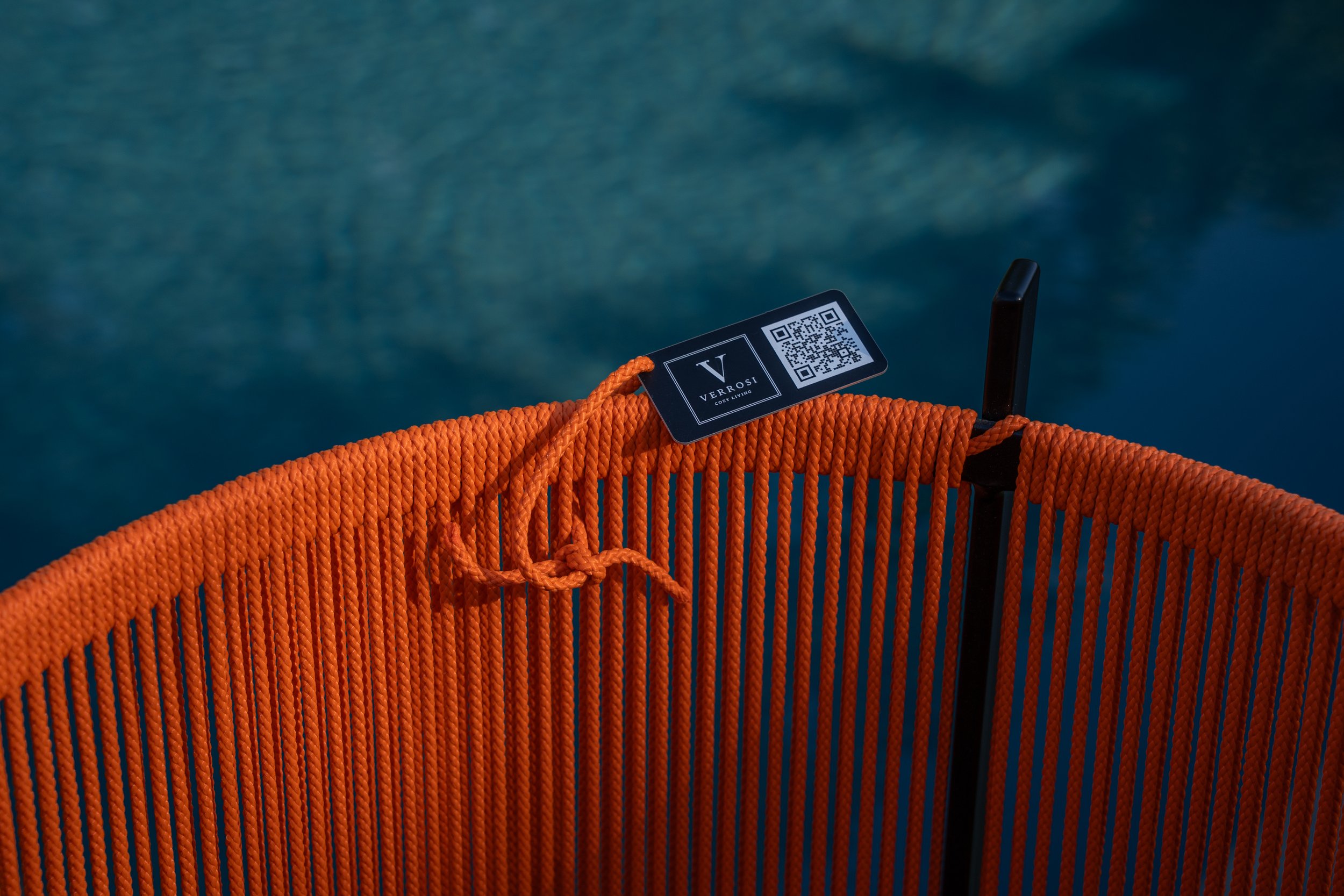 Close-up of an orange rope basket with a black handle, with a tag attached that reads 'VERROSI' and has a QR code, over a blurred blue-green background.