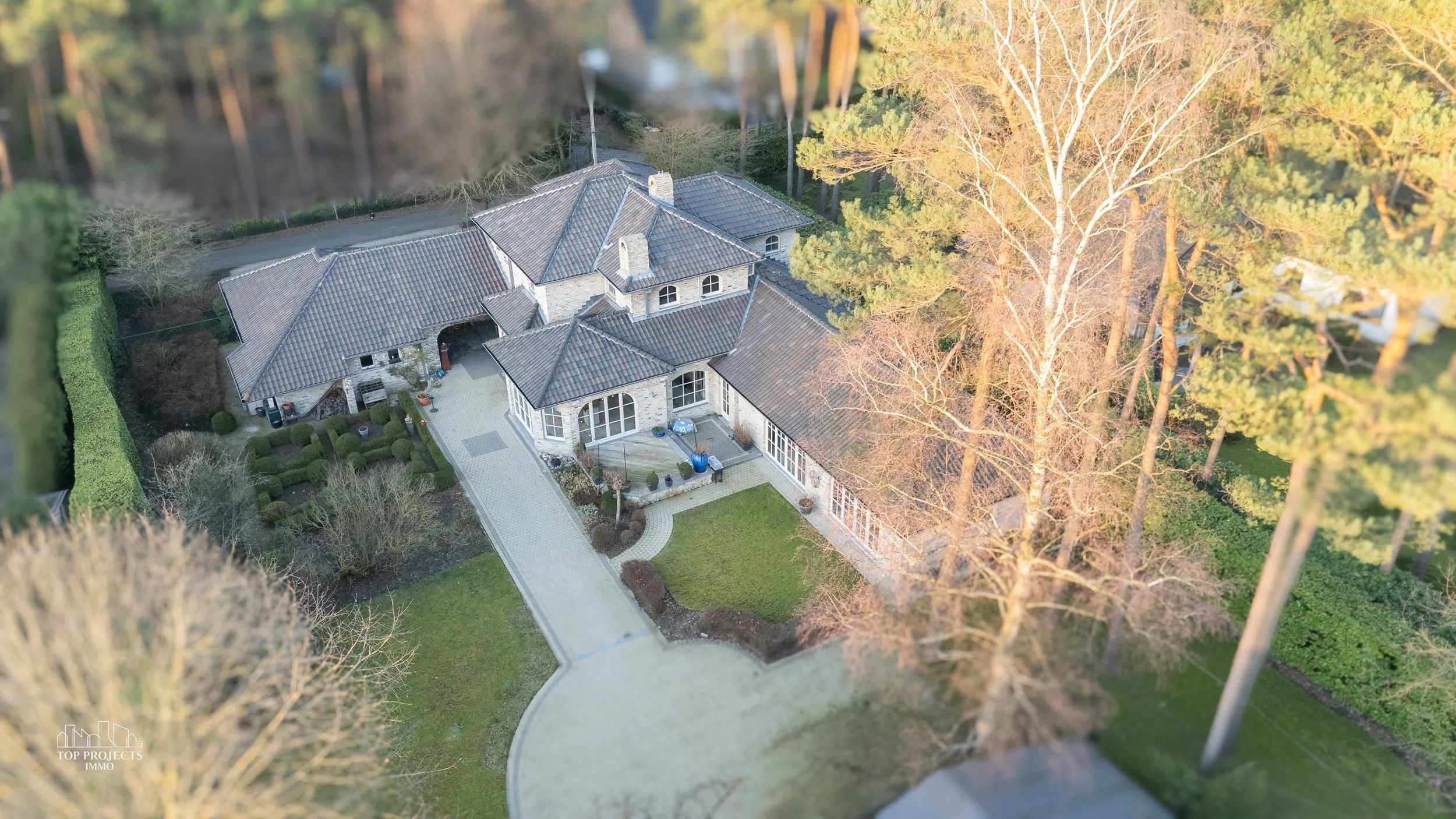 Aerial view of a large house with gray tiled roof, surrounded by trees and a green lawn, with a curved driveway leading to the entrance.