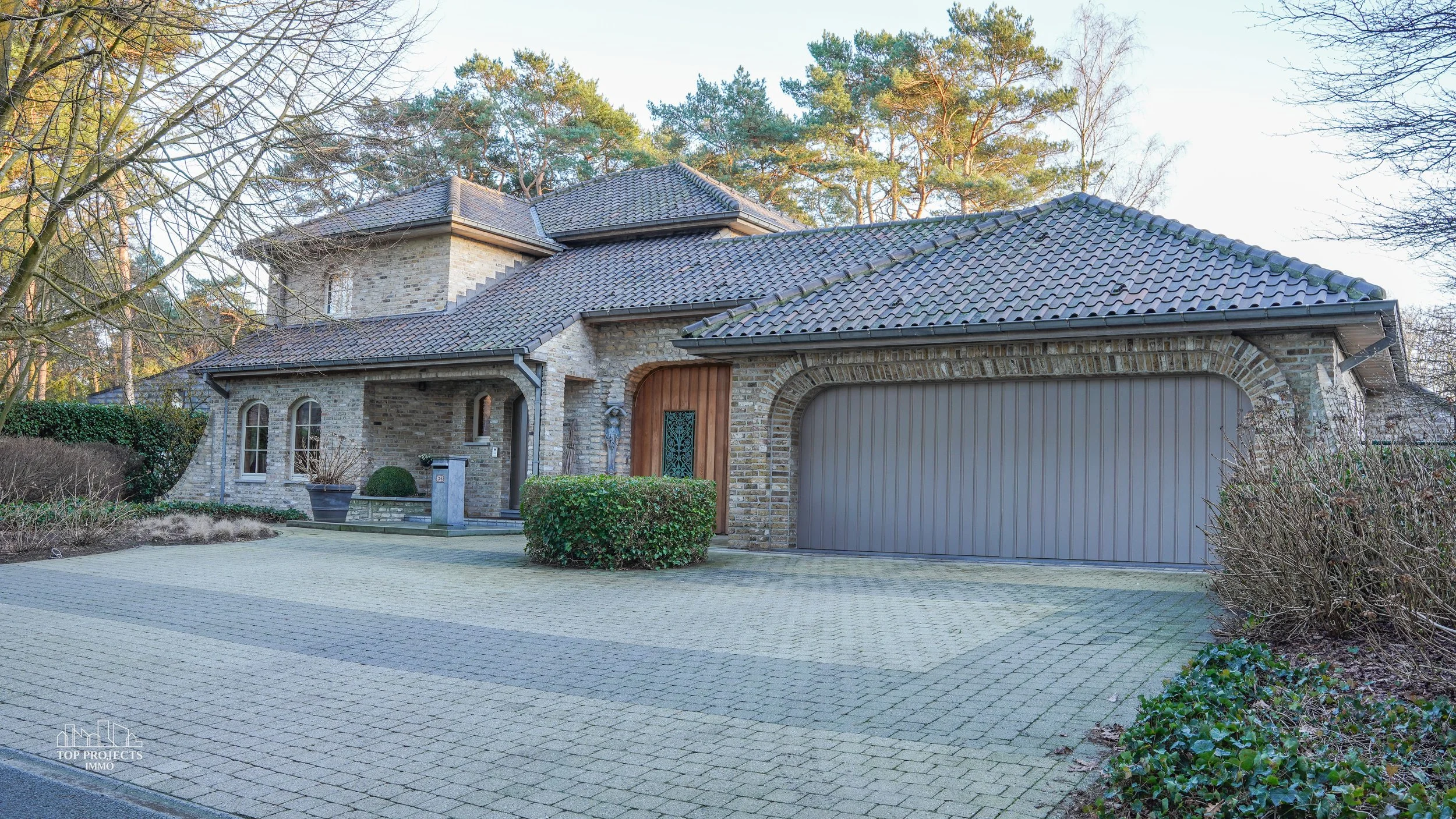 A brick house with a tiled roof and two garage doors, surrounded by trees and a paved driveway.