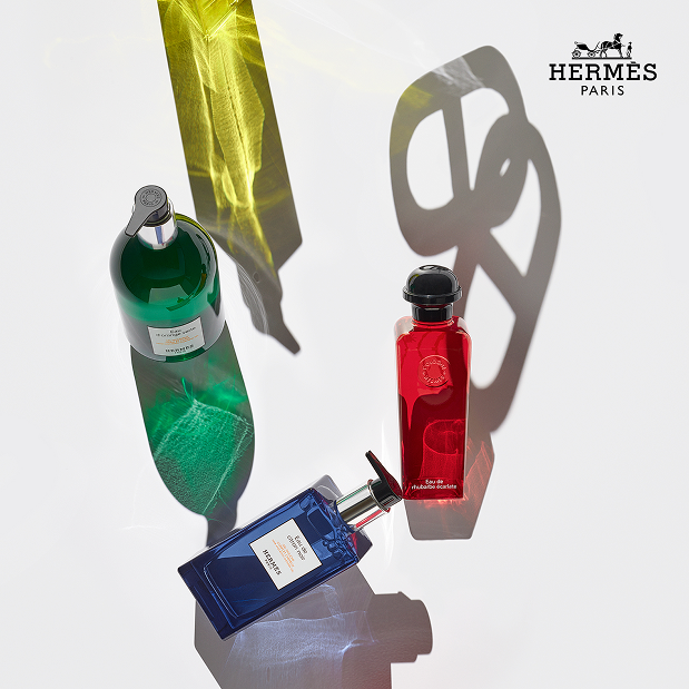 anne bassam - exécution graphique Trois flacons de parfum Hermès posés sur une surface blanche, avec des ombres colorées et le logo Hermès Paris en haut à droite.