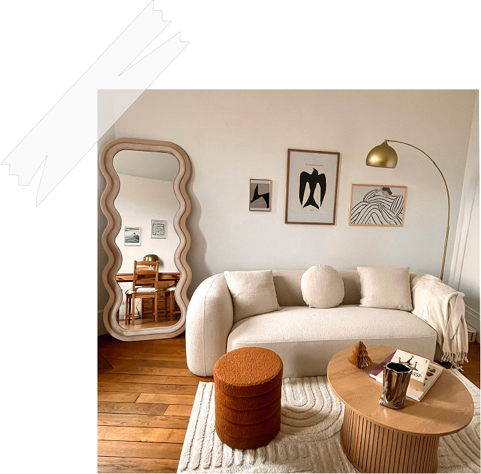 Anne Bassam - Salon moderne avec canapé beige, table basse en bois, étagère en miroir incurvée, tableaux abstraits, lampadaire en métal doré, tapis rayé blanc et beige, murs blancs, planche à manger en bois visible dans la pièce voisine.