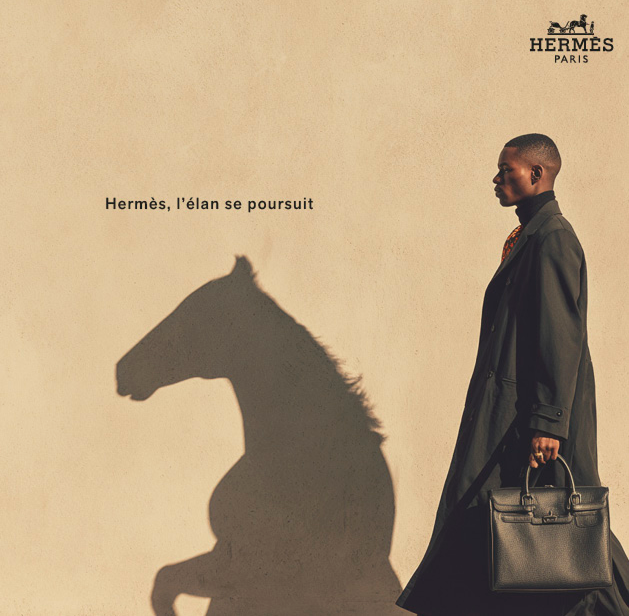 anne bassam - visuel généré pour campagne hermès fictive via midjourney Une personne en manteau noir marche devant un mur beige, créant une ombre en forme de cheval. Logo Hermès en haut à droite et texte "Hermès, l'élan se poursuit" à gauche.