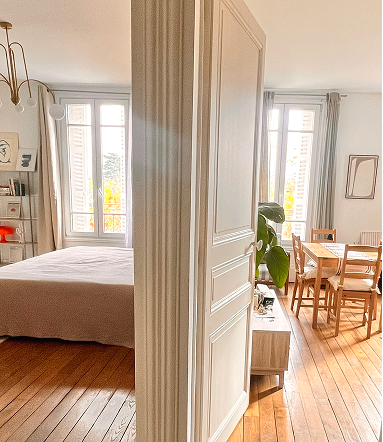 Anne Bassam - Une vue d'un appartement avec une chambre à gauche et une salle à manger à droite, séparées par une porte entrouverte.