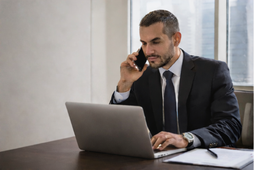 Businessman in a suit sitting at a desk, talking on a cellphone, using a laptop in an office.