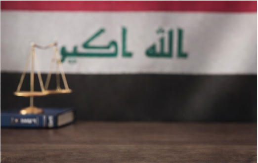 Miniature gold balance scale, two blue books labeled 'SAMPLE,' and an Iraqi flag in the background.