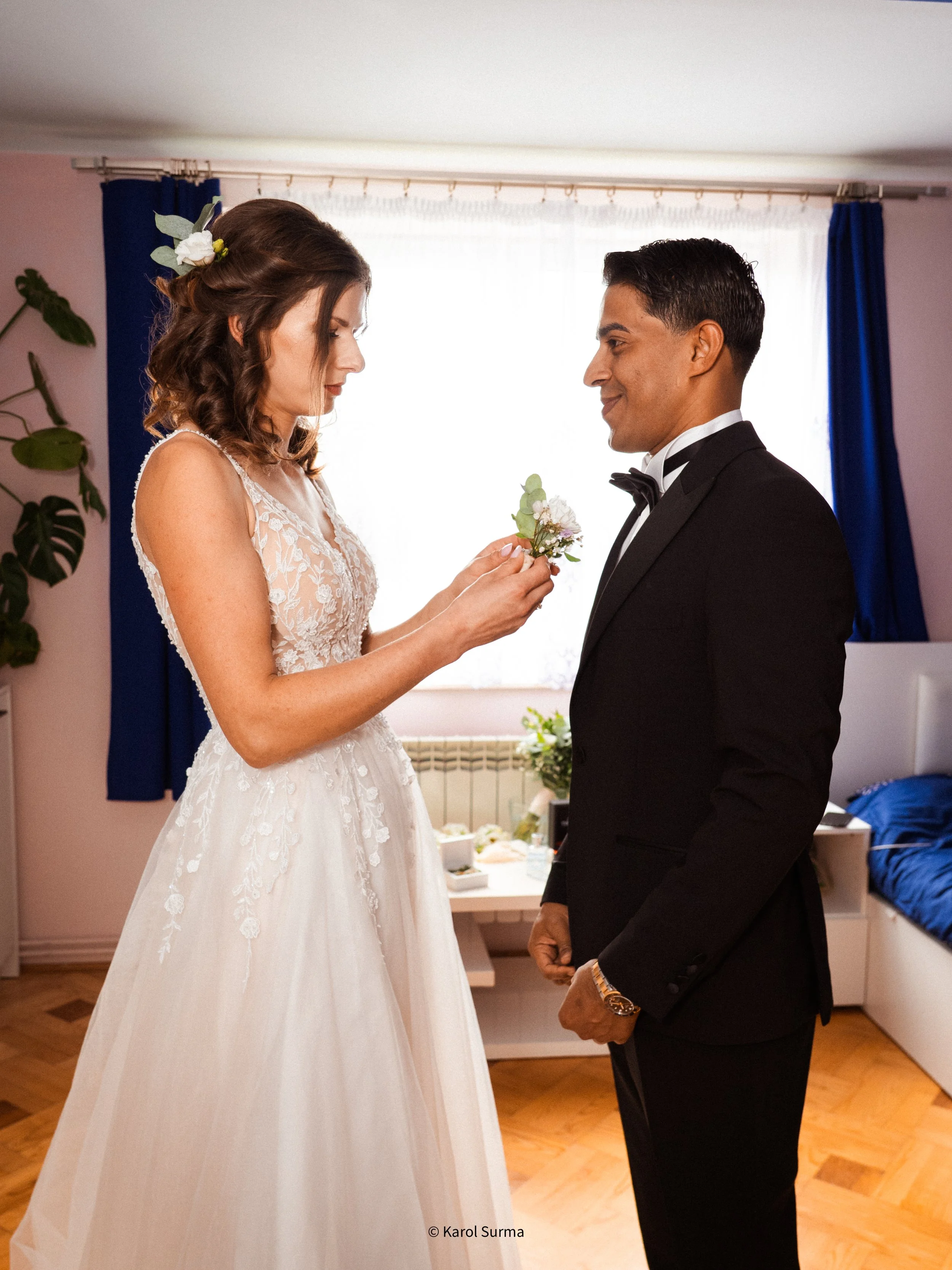 A bride in a white lace wedding dress puts a boutonniere on the groom, who is dressed in a black tuxedo, in a room with blue curtains and windows with white drapes.