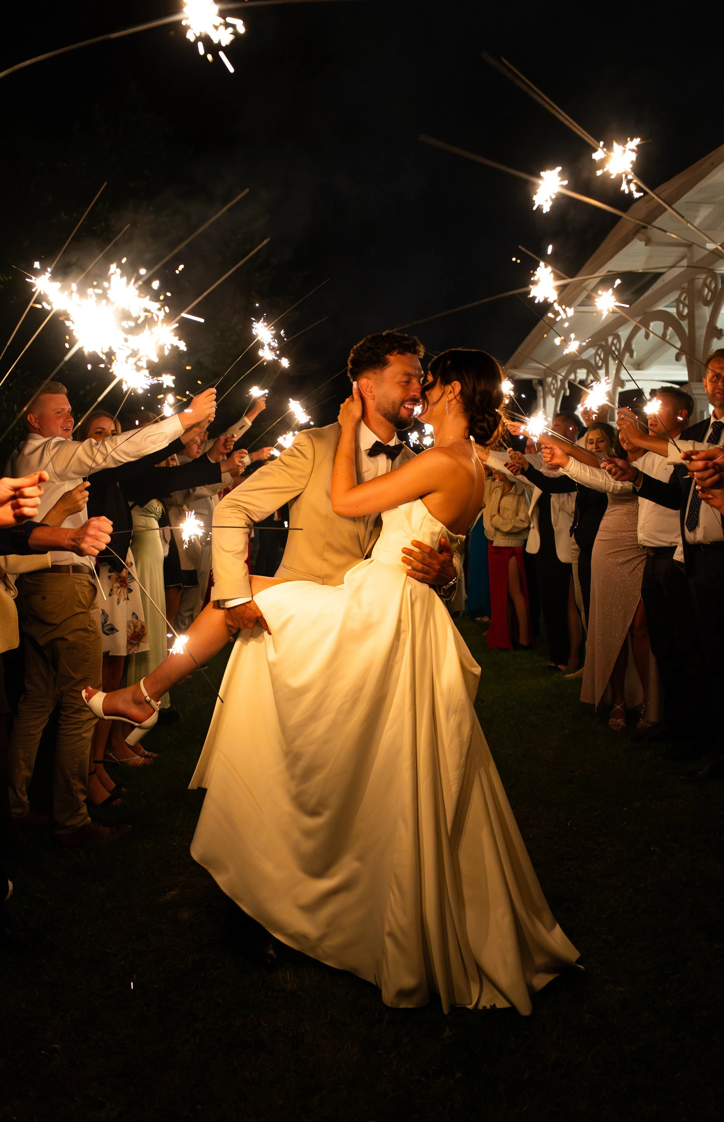 A newlywed couple dancing among friends holding sparklers at night during a wedding celebration.