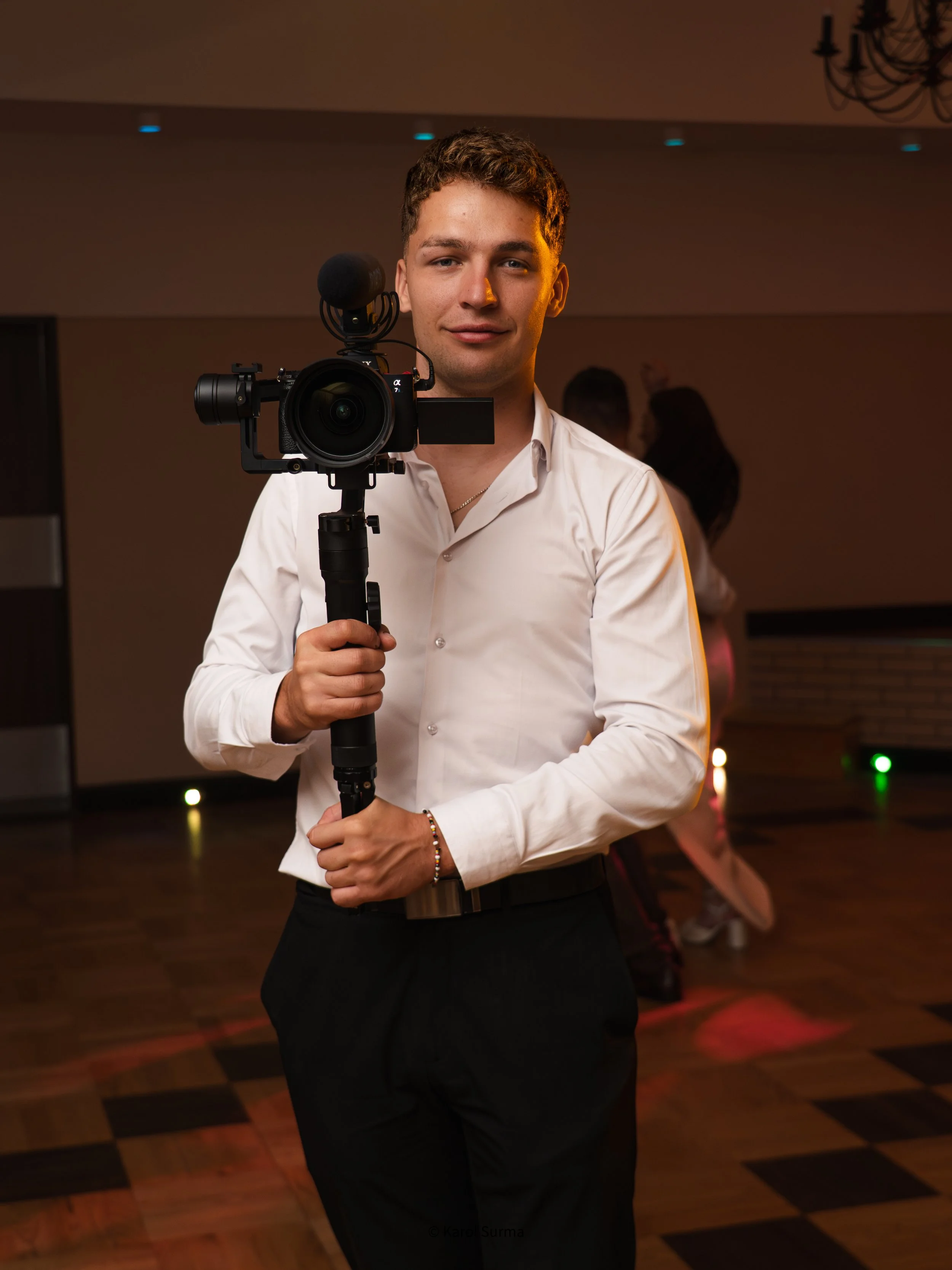 Young man in white shirt holding a professional camera on a stabilizer at an indoor event.