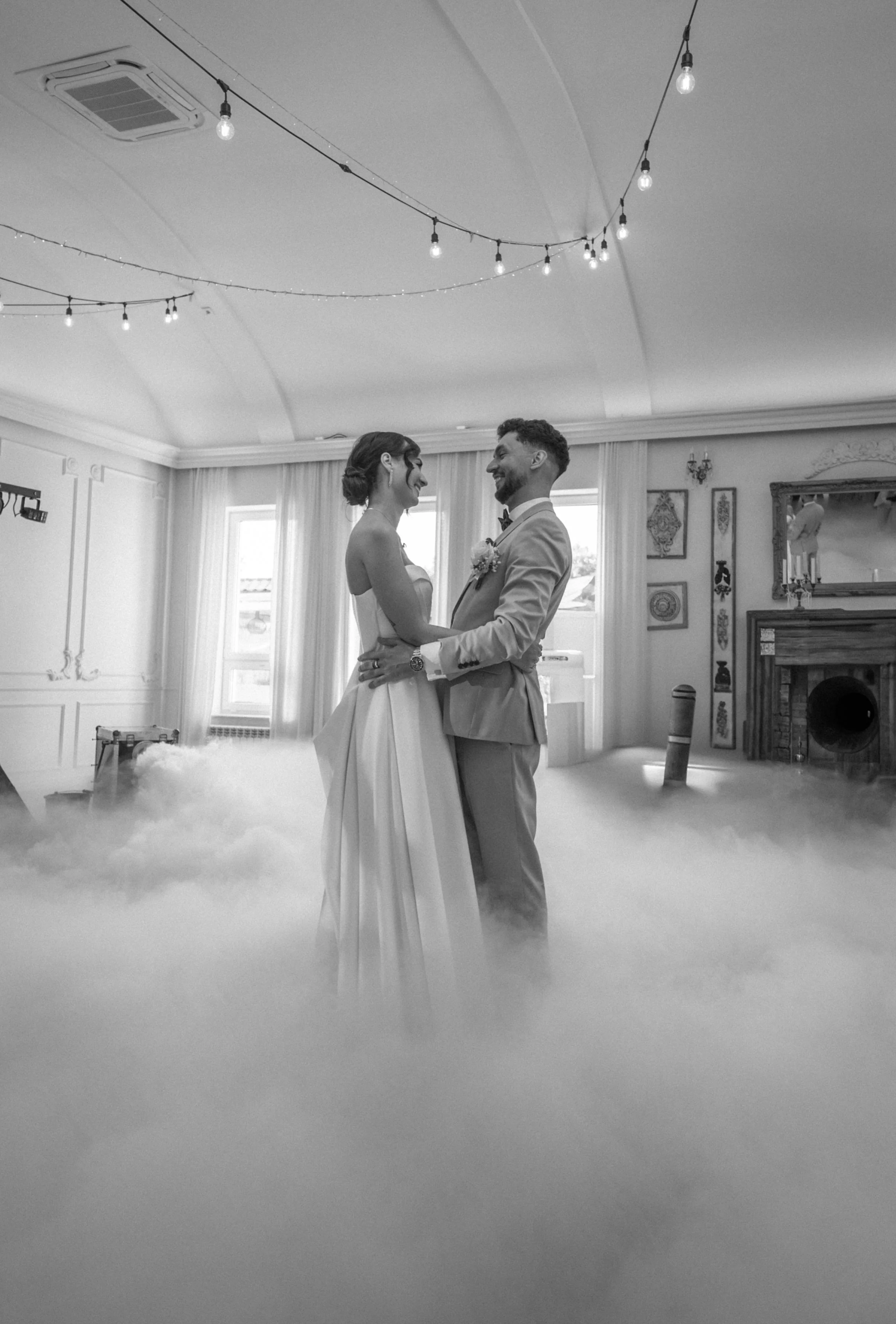 A bride and groom dancing in a wedding reception hall, surrounded by a cloud of fog, with string lights hanging from the ceiling.