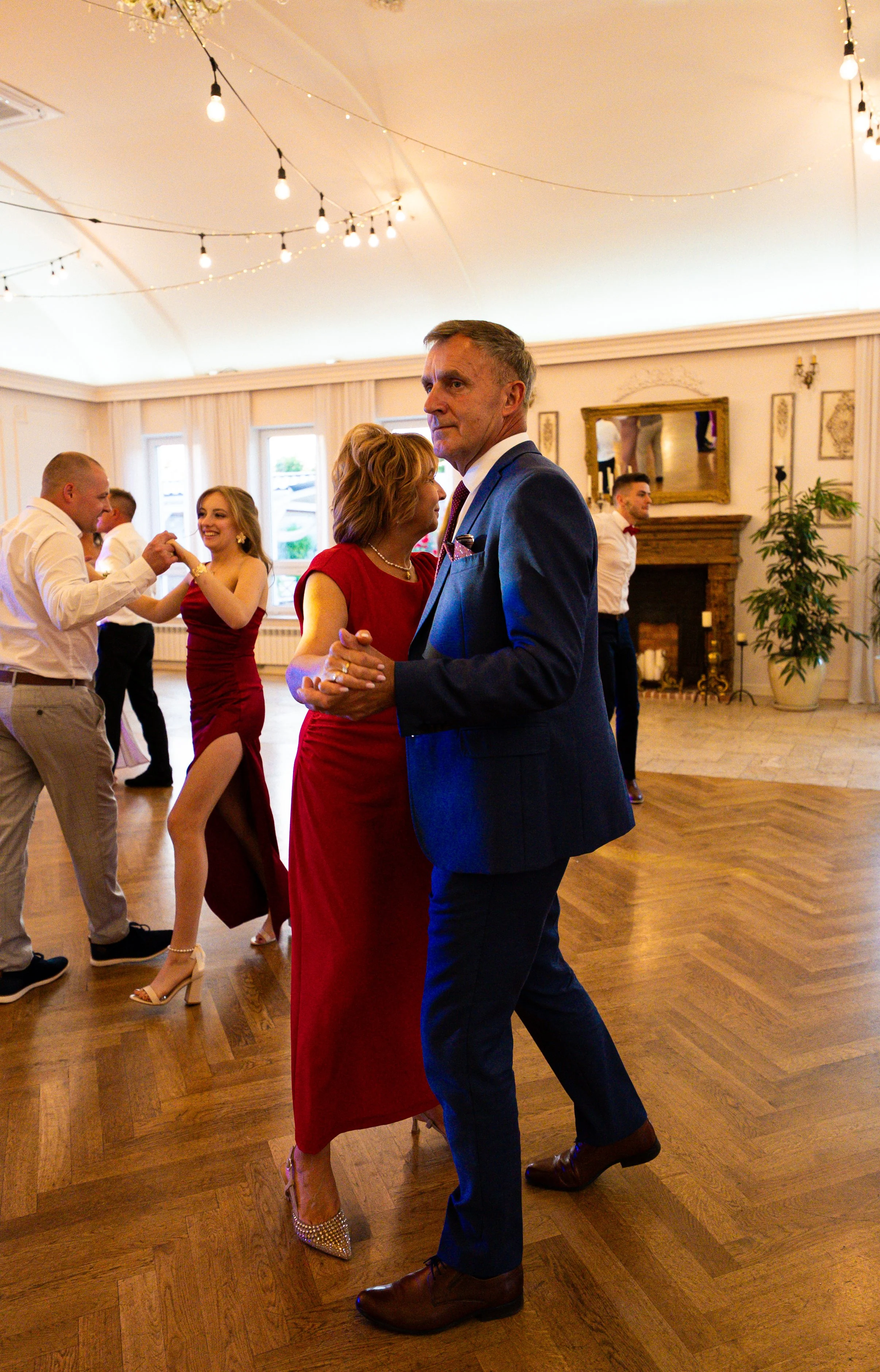 People dancing at a celebration or wedding in a decorated hall with string lights, a fireplace, and elegant decor.