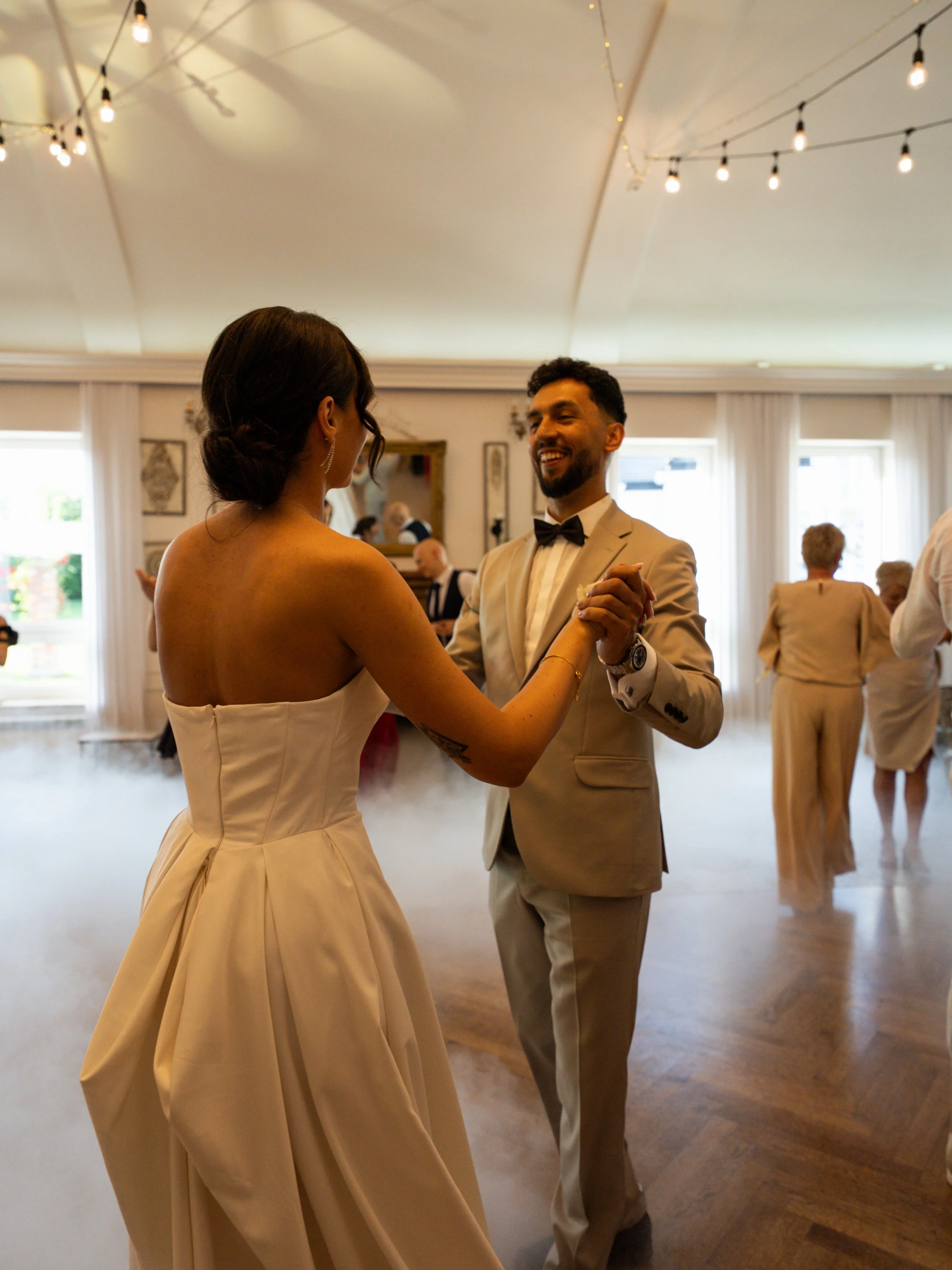 A couple dancing at a wedding reception, with string lights overhead and fog on the dance floor, in a decorated indoor venue.