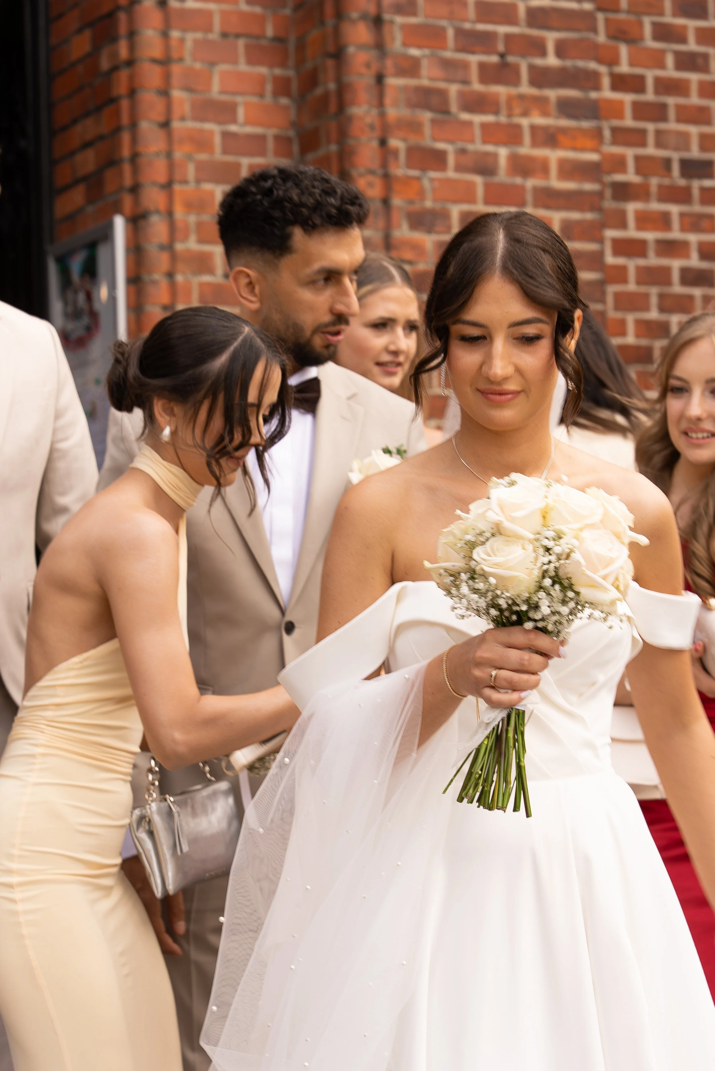 A bride holding a bouquet of white roses, surrounded by wedding guests dressed in formal attire, outside a brick building.
