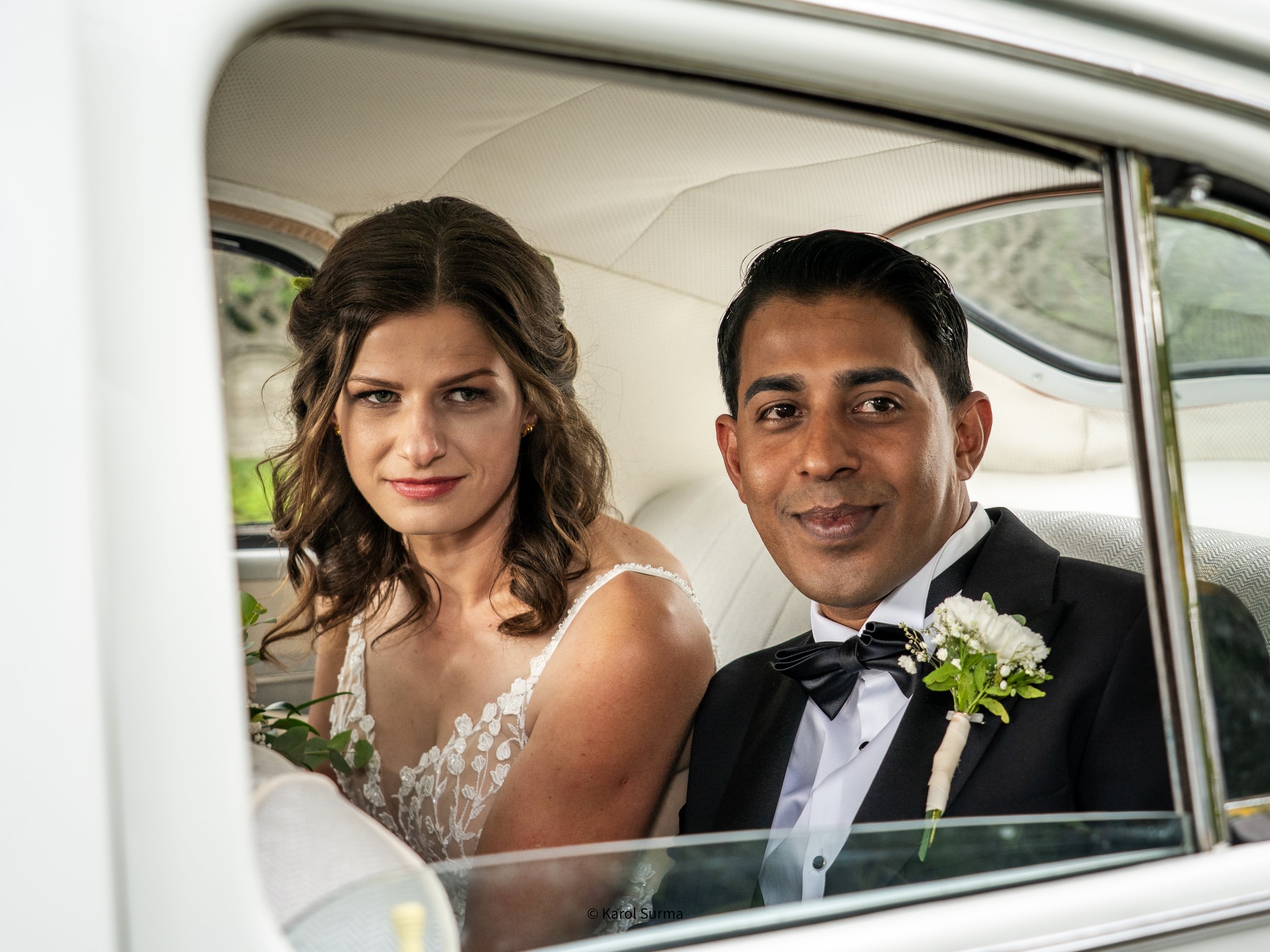 A bride and groom sitting in the backseat of a vintage white car, smiling.
