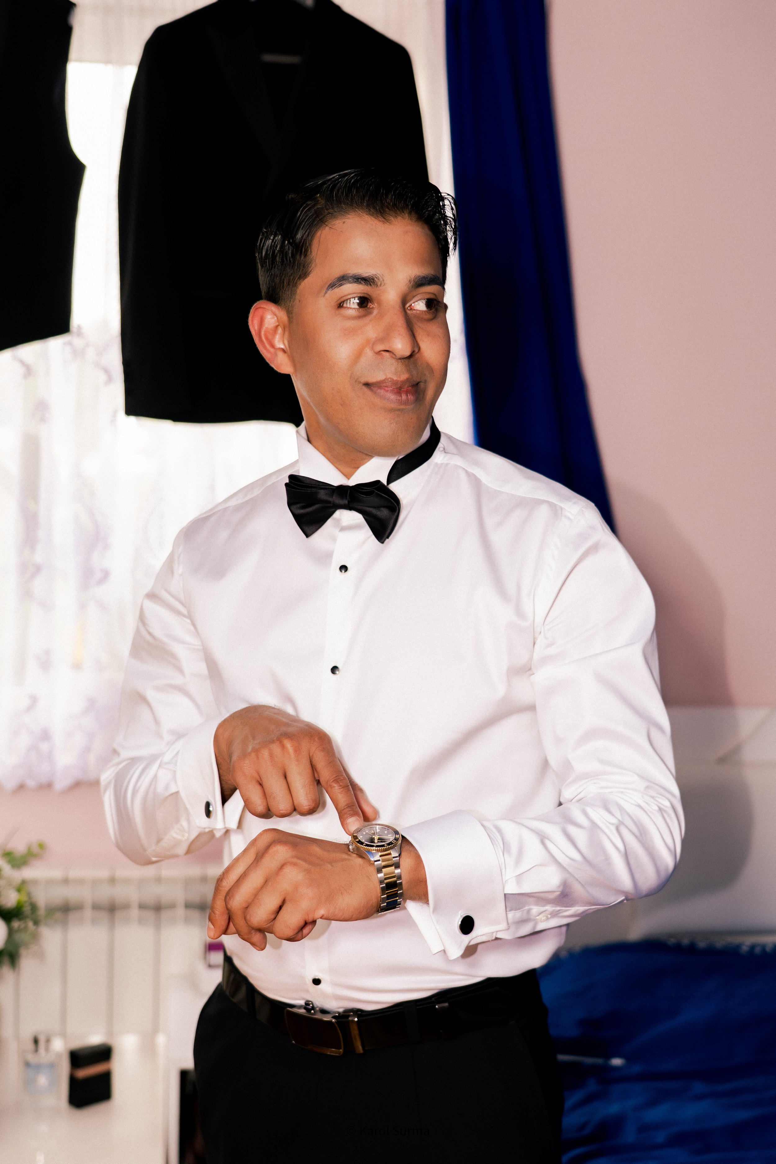 A man dressed in a white shirt with a black bow tie, wearing a watch, looks at his wrist while preparing for a formal event in a room with a black suit hanging in the background.